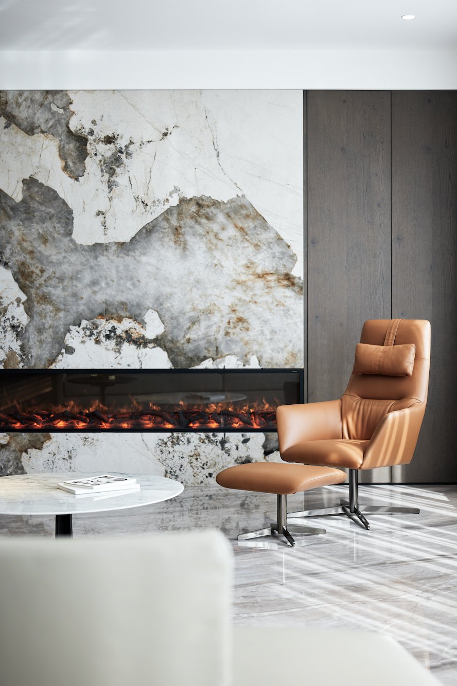 Modern living room with tan leather chair, marble wall with fireplace, and round white table.