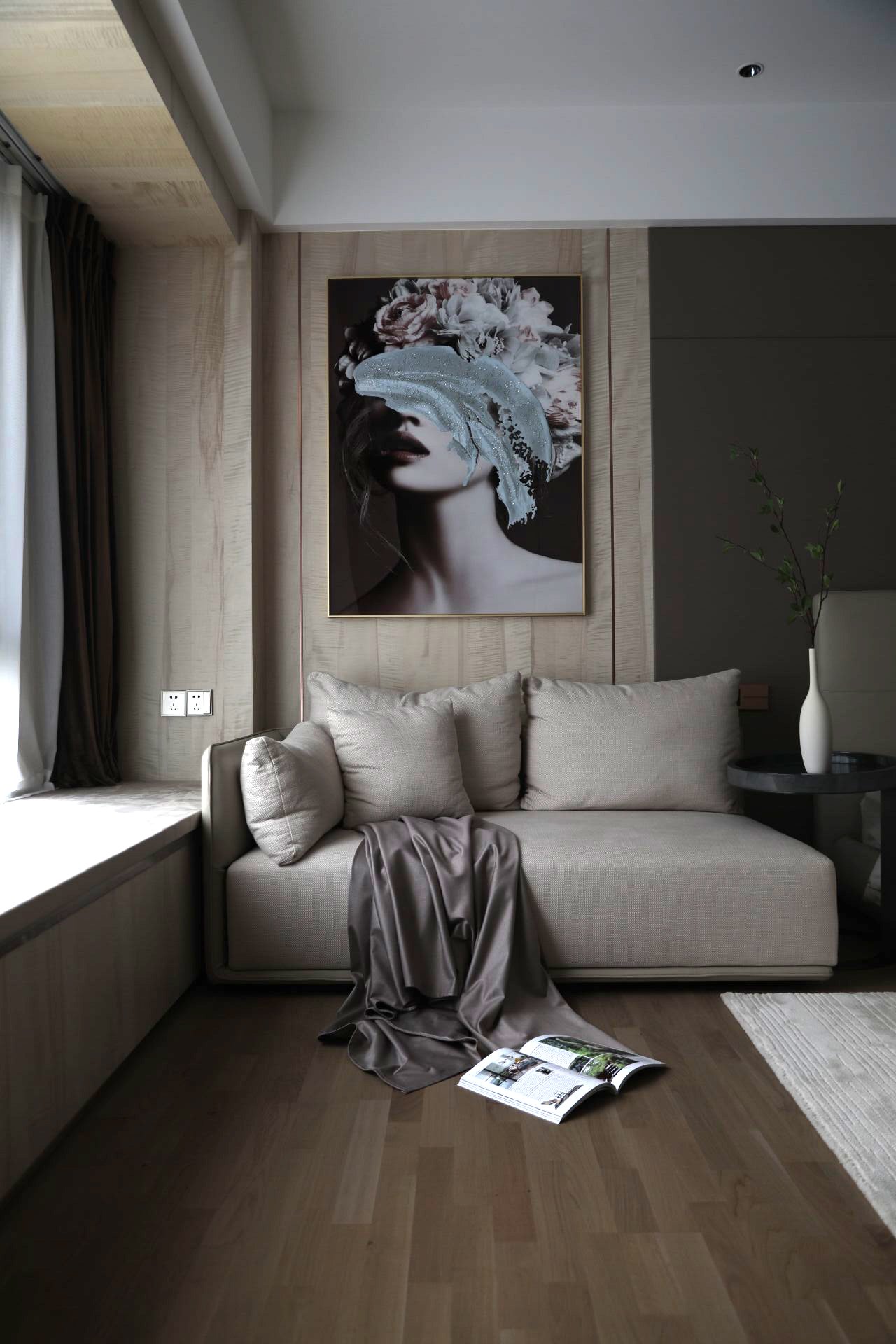 Modern living room with beige sofa, artistic wall portrait, soft draped fabric, open magazine on the wooden floor, and vase on side table.