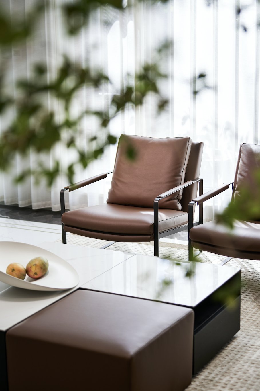 Modern living room with brown leather chairs, white curtains, and a coffee table featuring a bowl with two mangoes.