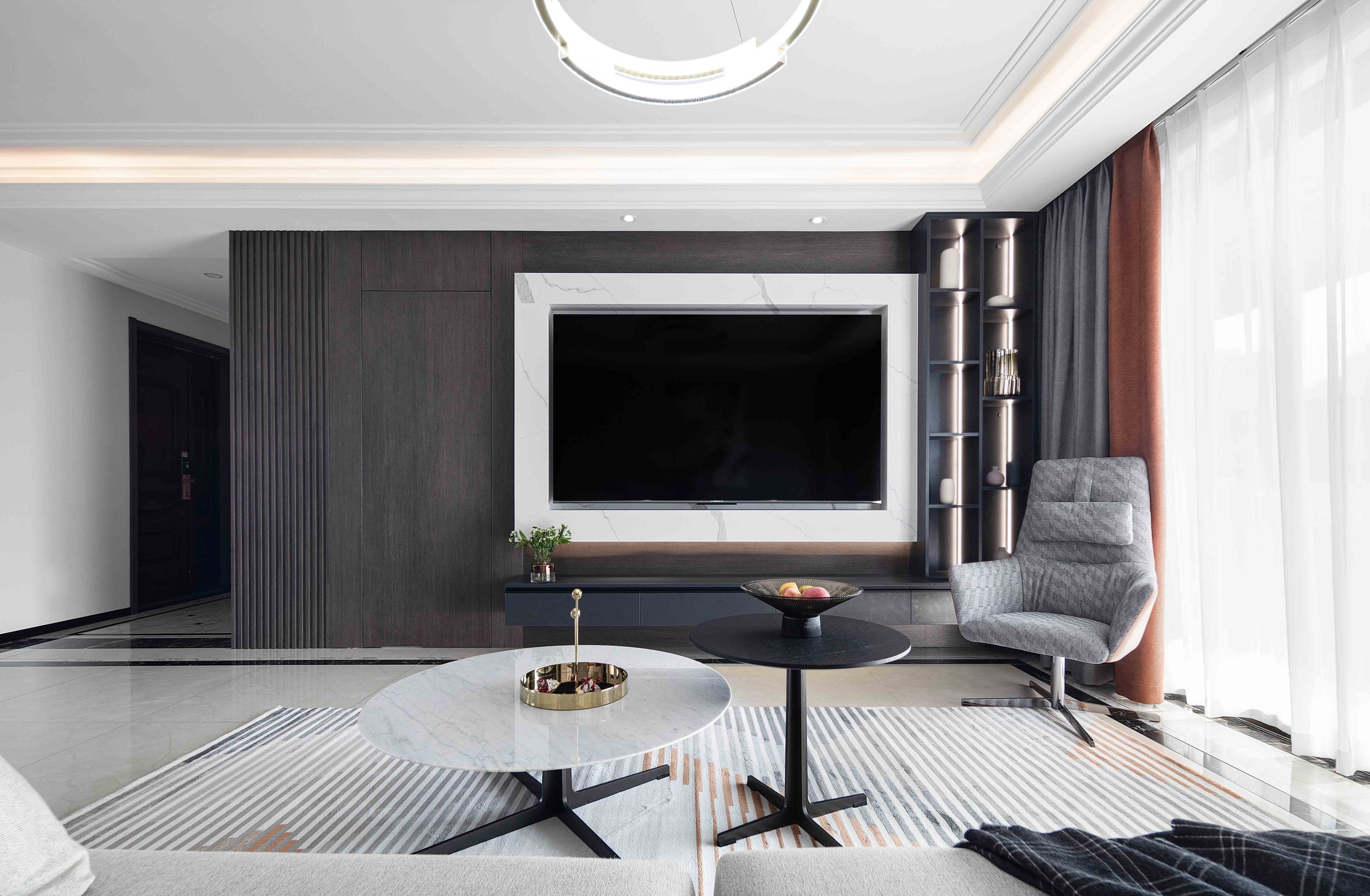 Modern living room with a large television on a dark accent wall, a gray armchair, a round marble coffee table, and decorative items on side tables, with sheer curtains and stylish lighting.