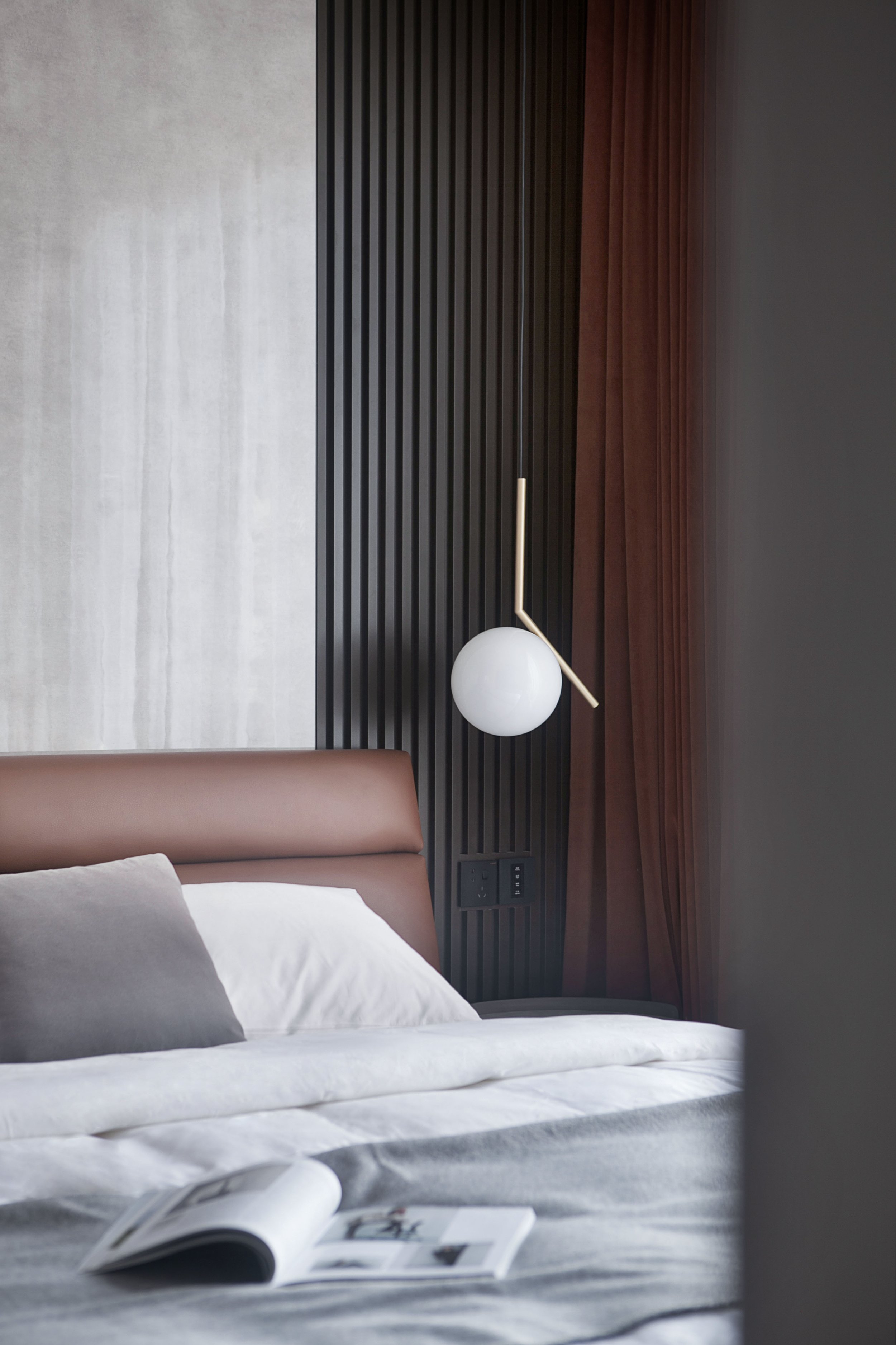 Modern bedroom interior with a brown leather headboard, white bedding, gray blanket, and a gray pillow. A stylish wall lamp is mounted on a black paneled wall. An open book is on the bed, and brown curtains are visible.