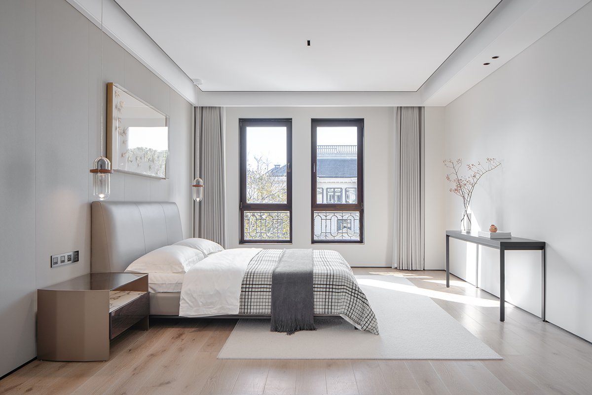 Minimalist bedroom with a bed, modern pendant lights, two large windows, and a narrow console table adorned with a vase and books.