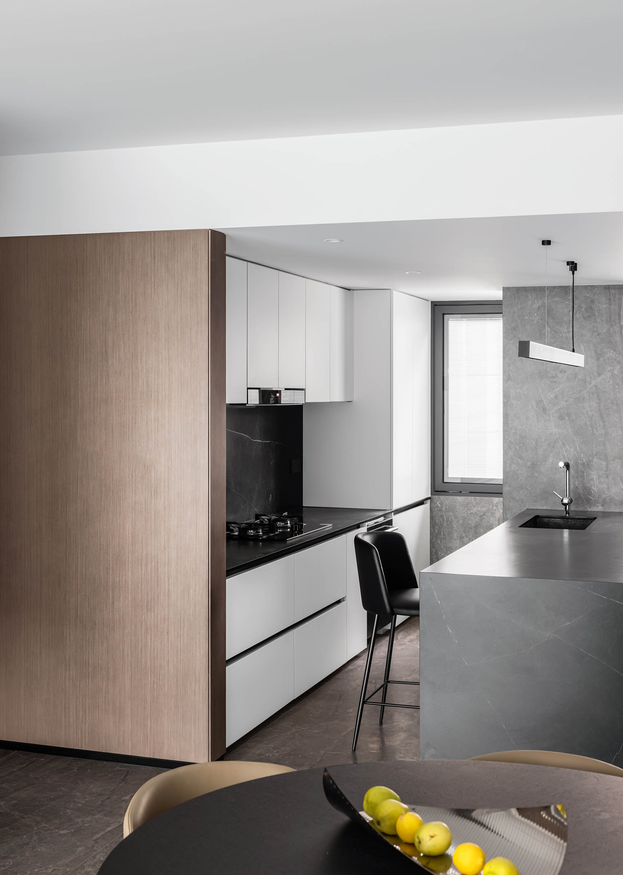 Modern kitchen with sleek gray countertops, black bar stool, white cabinets, and a gas stove. A minimalist pendant light hangs above the sink area. A window with blinds provides natural light. A table with a fruit bowl is in the foreground.