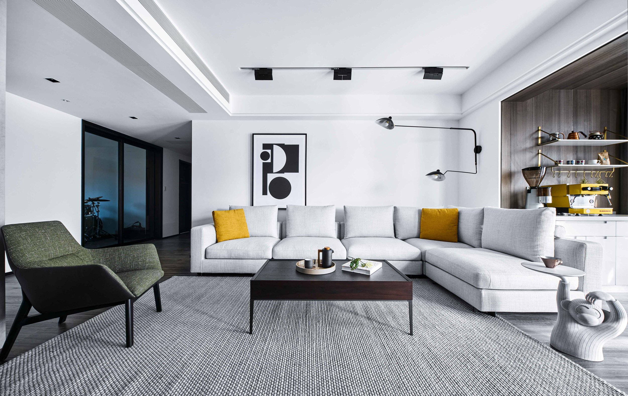 Modern living room with a gray sectional sofa, yellow accent pillows, and a dark wood coffee table on a textured rug. A green armchair and a small side table with a monkey-shaped base are also present. Wall art and a coffee station with shelving are