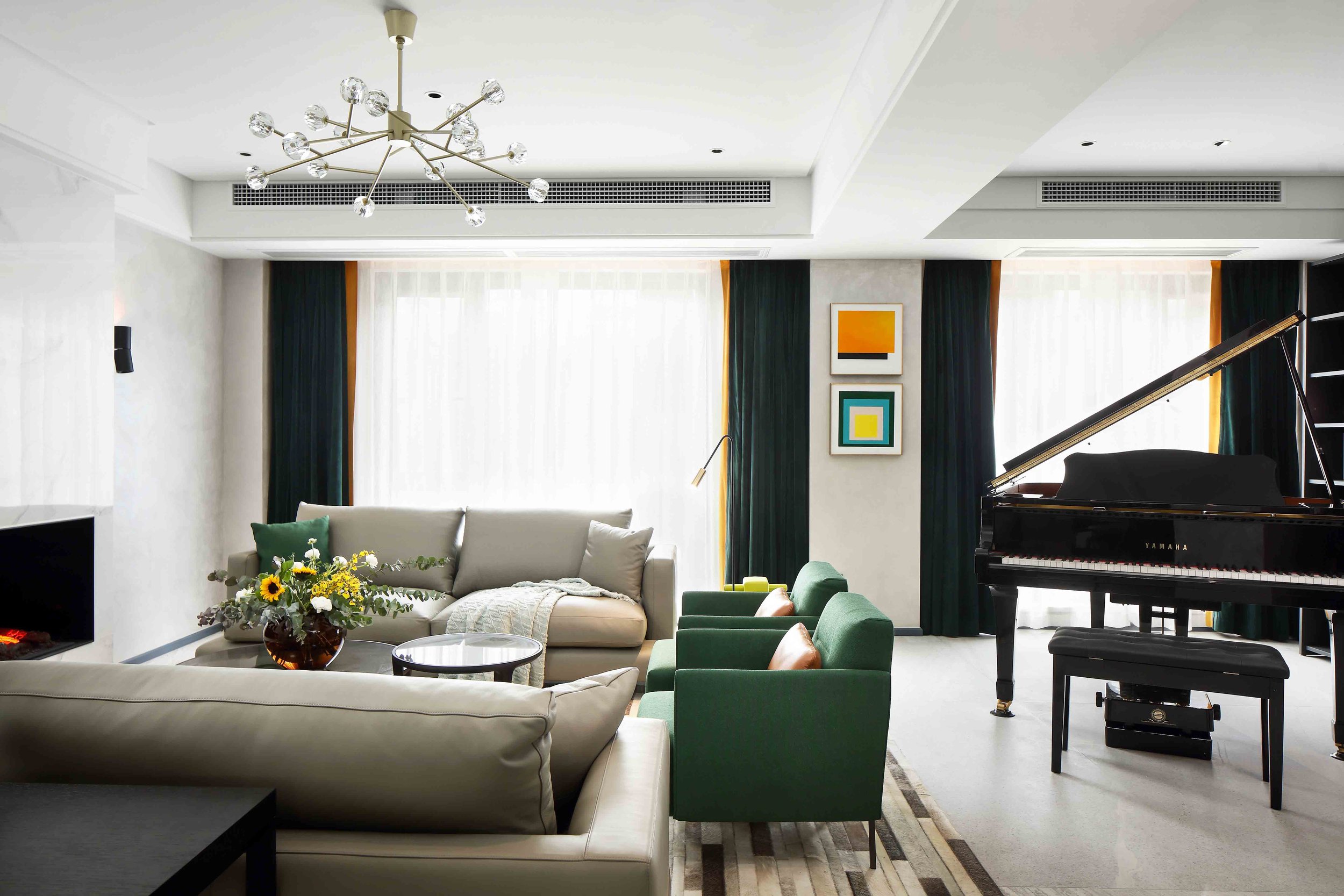 Modern living room with a beige sectional sofa, green armchairs, a grand piano, and a fireplace. A contemporary chandelier hangs from the ceiling. Two abstract paintings are on the wall, and a vase of flowers is on the coffee table.