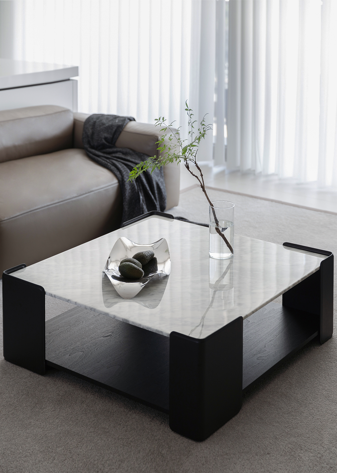 Modern living room with a light brown leather sofa, black throw blanket, marble-top coffee table, vase with a plant branch, and a decorative bowl with stones.