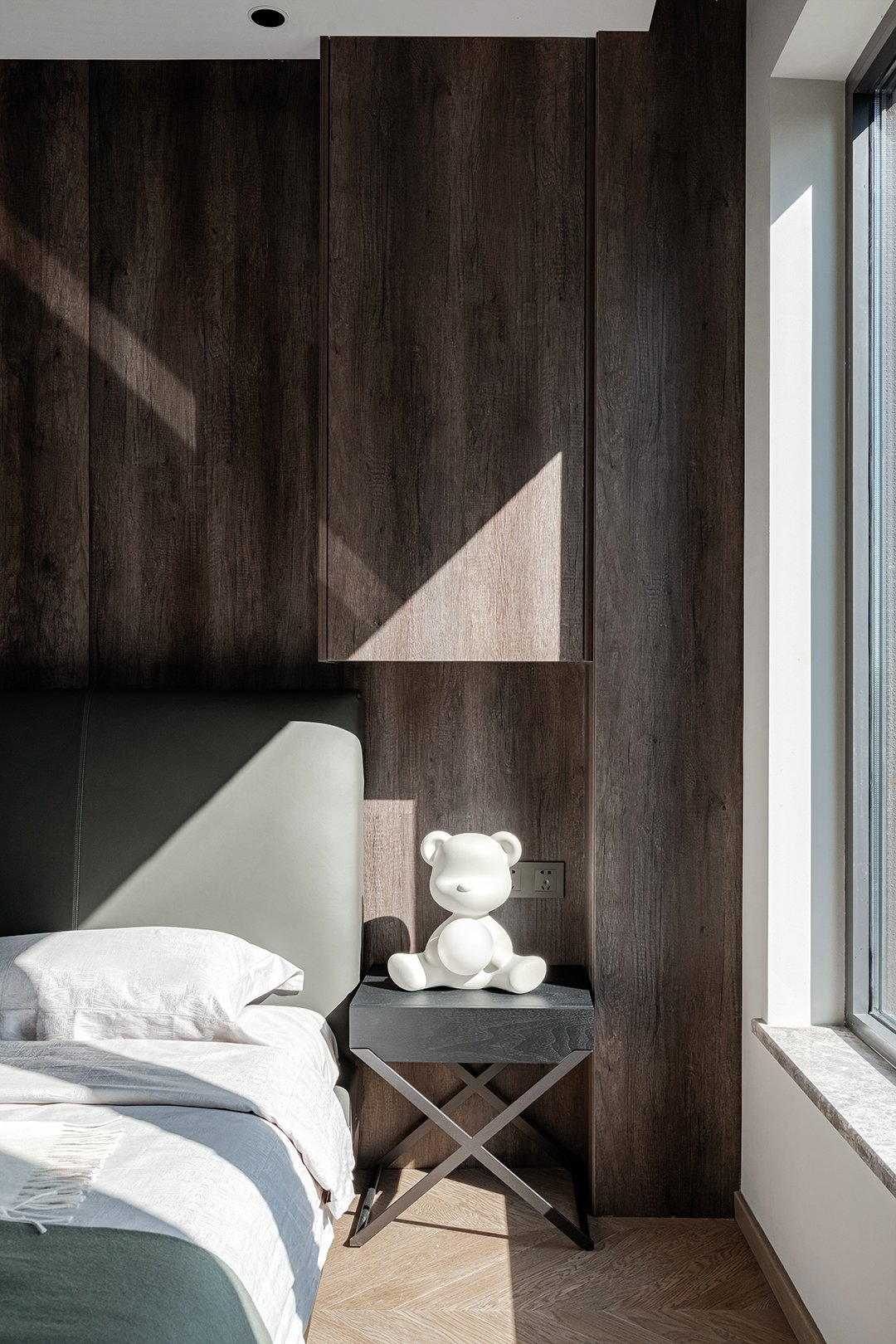 Bedroom with wooden wall panel, bed with white pillow and sheet, small side table with white teddy bear statue, and sunlight coming through a window.