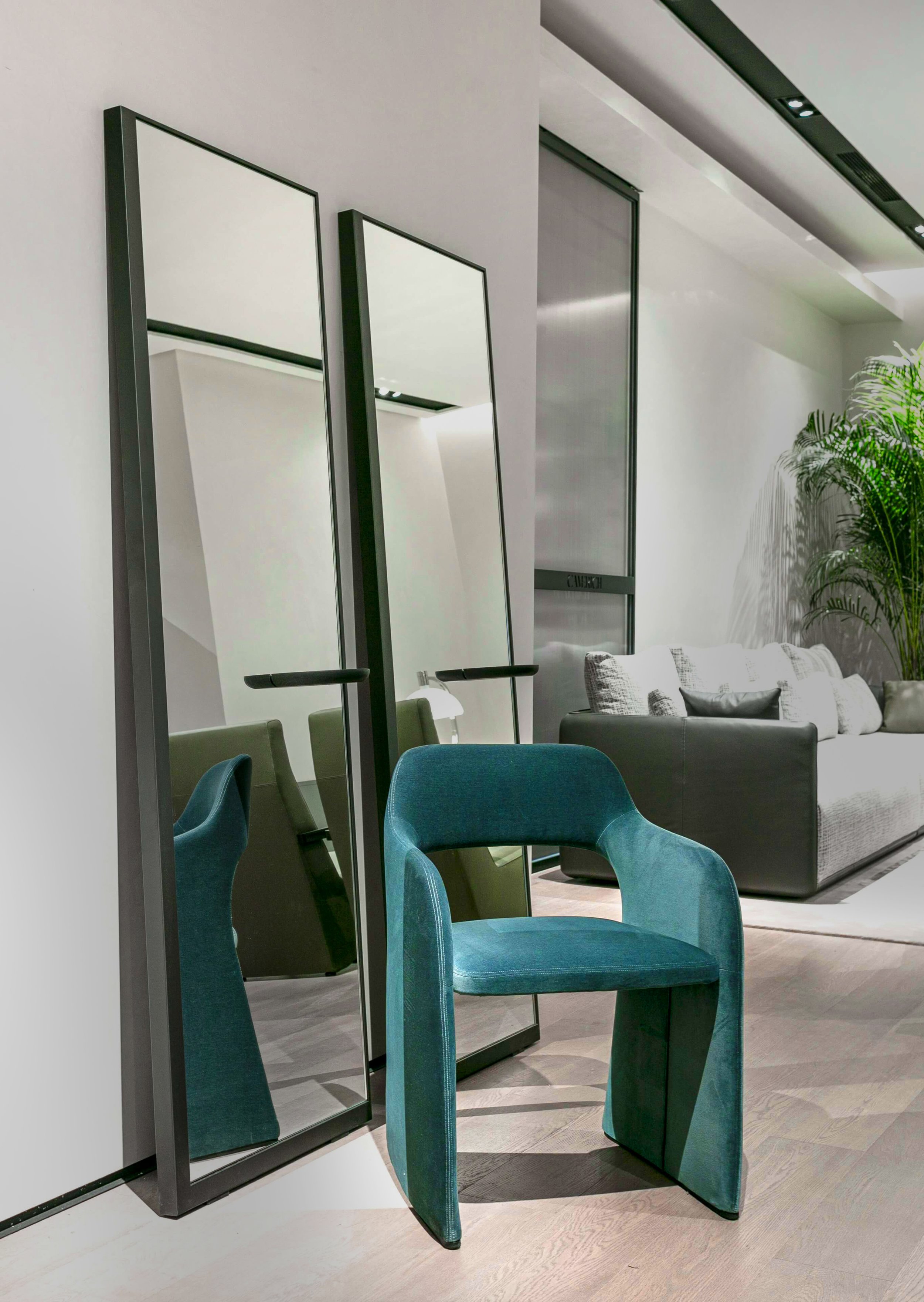 Modern interior with two tall mirrors, a teal chair, and a gray sofa, with green plants in the background.