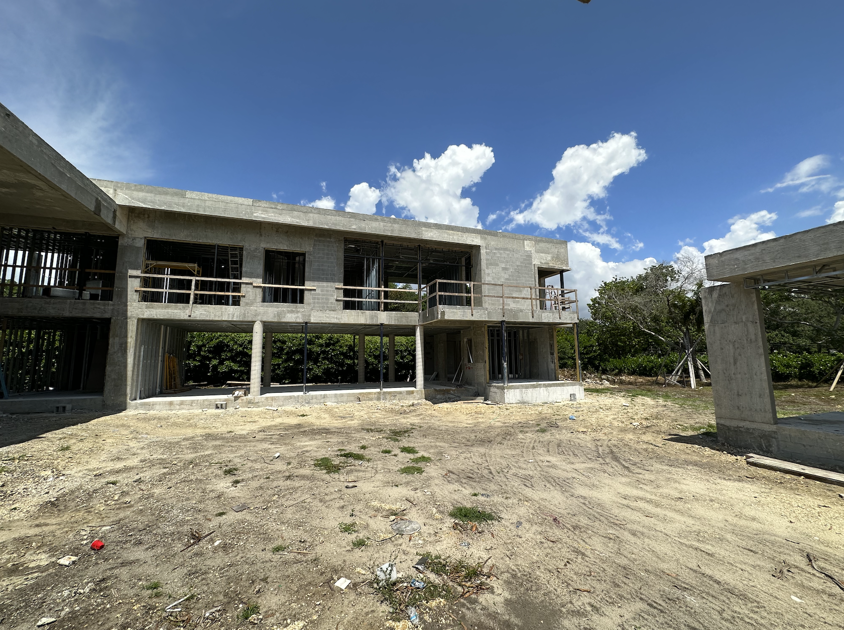 Under construction concrete building with two stories, surrounded by dirt and sparse vegetation under a clear blue sky with scattered clouds.