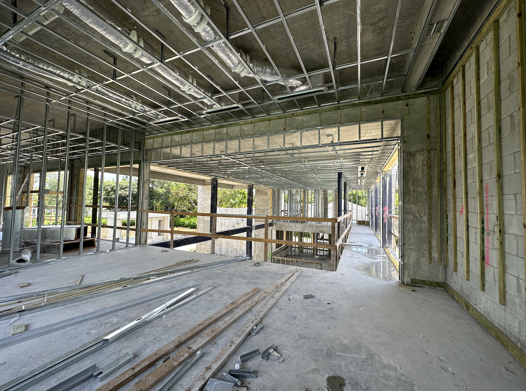 Interior view of a building under construction with exposed metal framing and ducts, unfinished walls, and a concrete floor. A walkway with wooden railings leads outside, offering a glimpse of greenery.