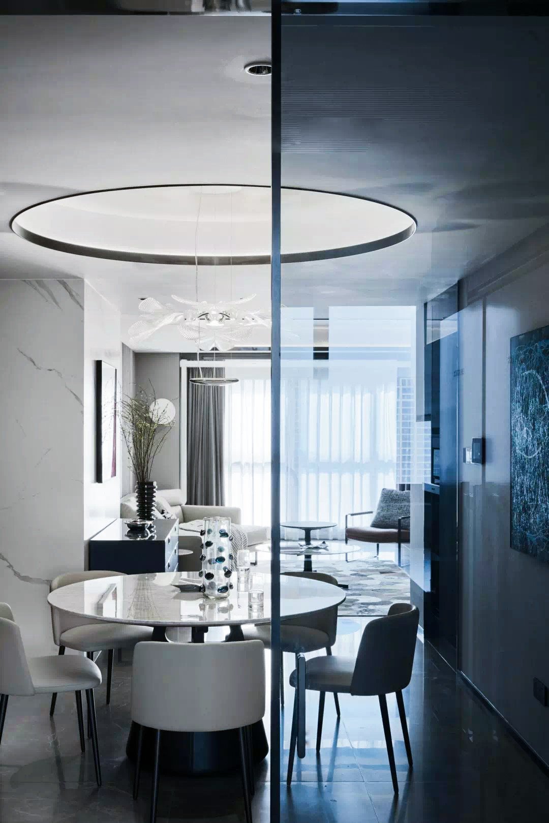 Modern dining and living area with a round table and chairs, contemporary chandelier, large windows with sheer curtains, and abstract wall art.