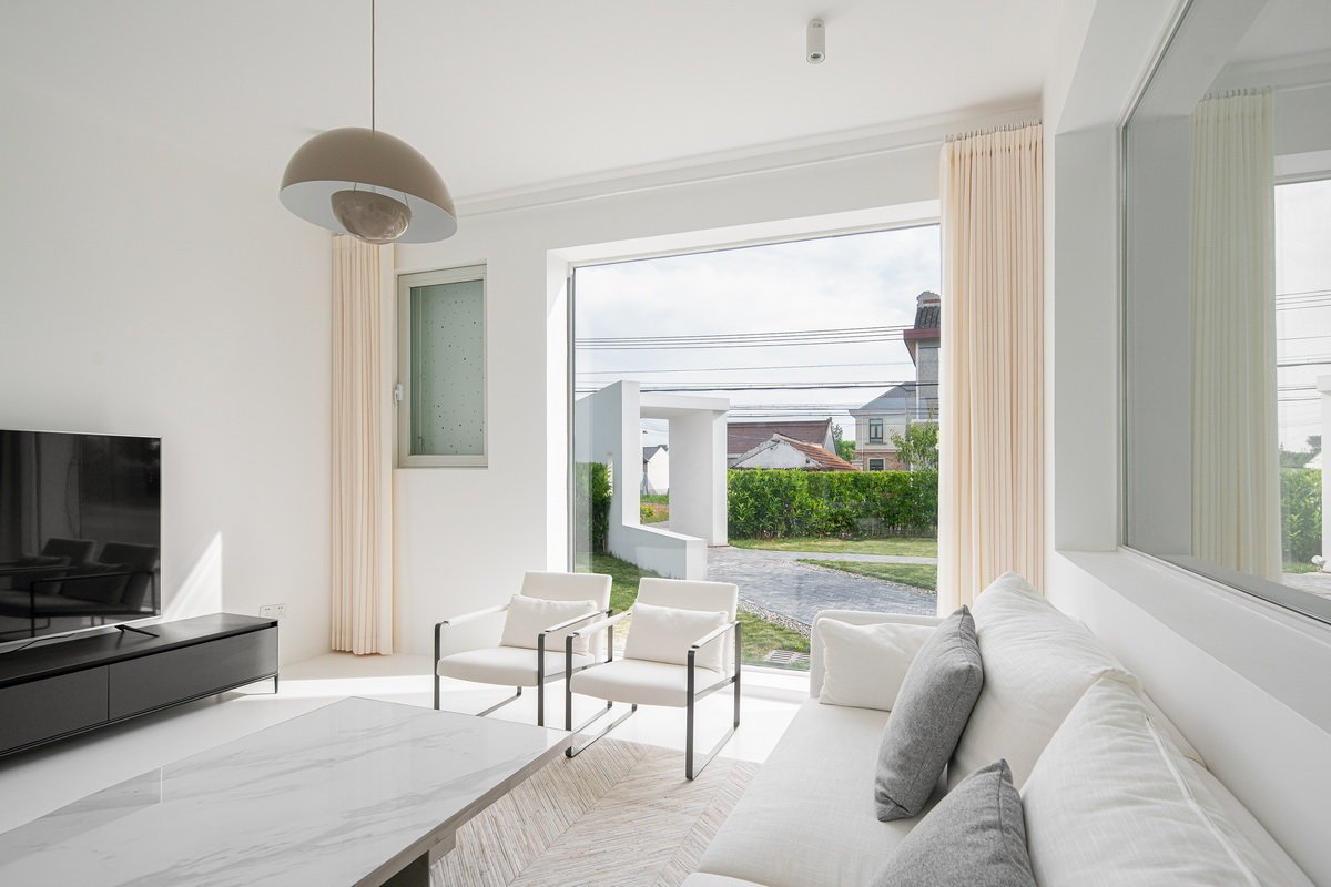 Modern living room with white sofa, armchairs, and TV; large window with view of driveway and house exterior.