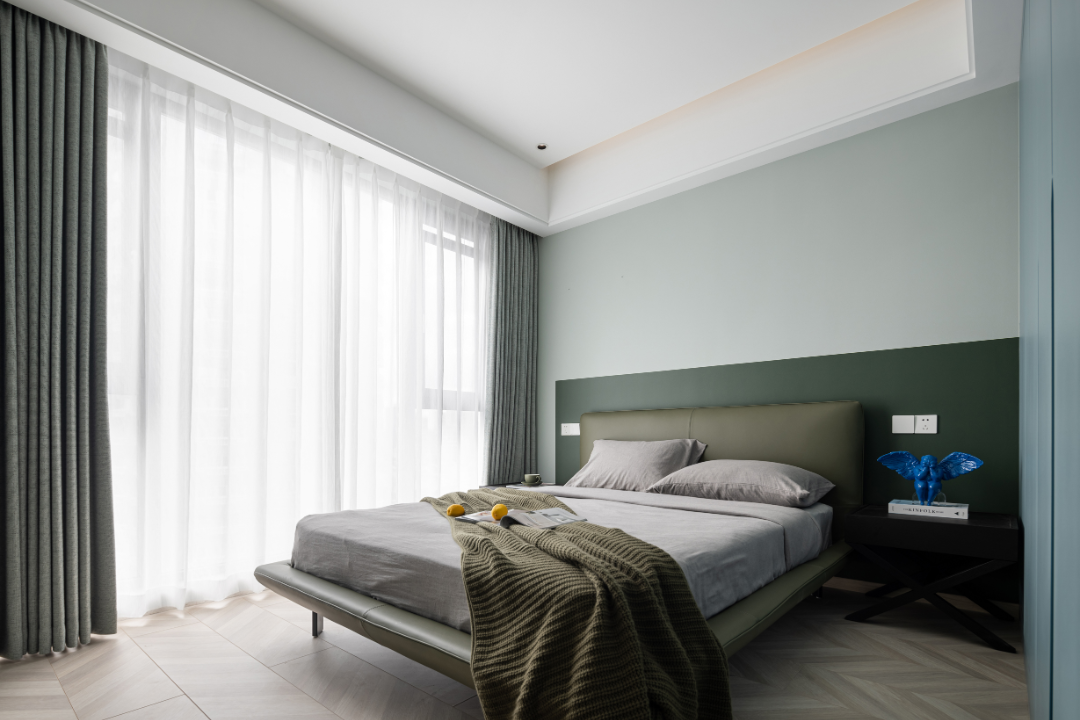 Minimalist bedroom with a modern bed, gray bedding, green accent wall, sheer curtains, and a side table with books and a blue sculpture.