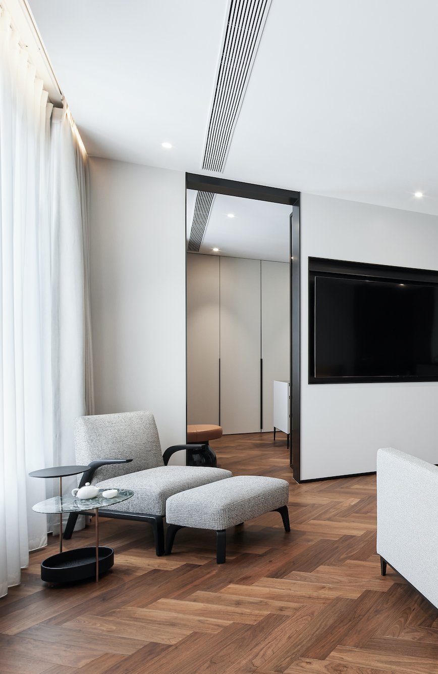Modern living room with a gray armchair and ottoman, wooden floor, small glass table, large flat-screen TV on the wall, and sheer white curtains.
