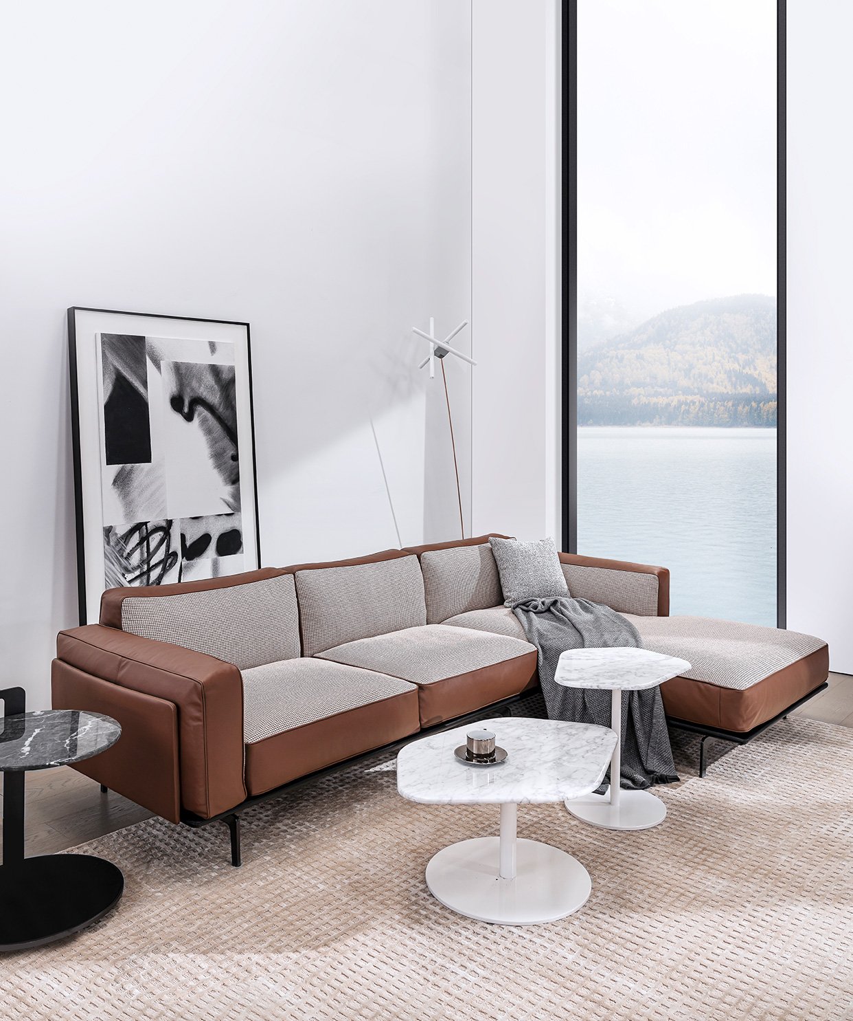 Modern living room with a brown and gray sectional sofa, abstract artwork, floor lamp, marble-top coffee tables, and a large window with a scenic lake view.