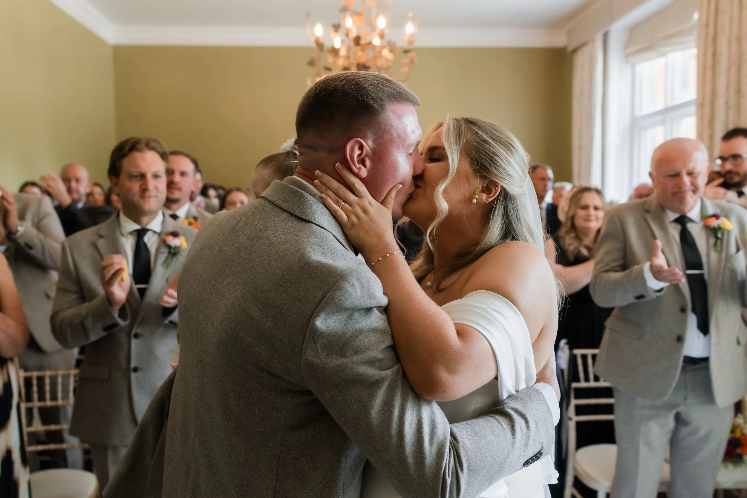 A bride and groom share a kiss during their wedding ceremony, surrounded by guests clapping and celebrating indoors with a chandelier and large windows.