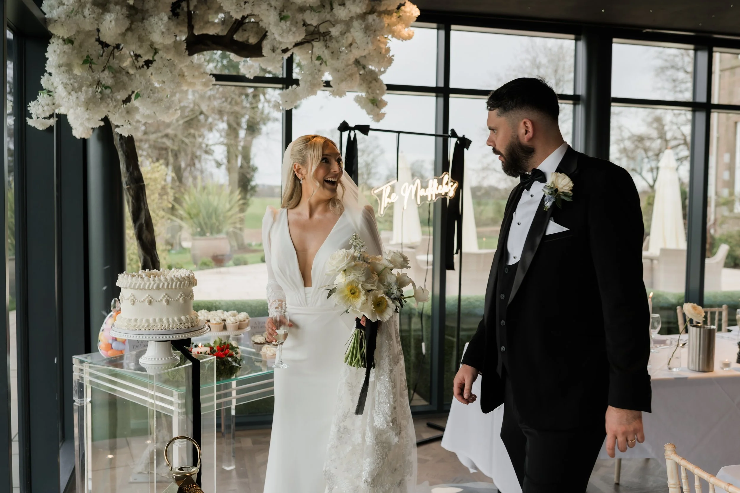 A bride and groom are at their wedding reception, standing near a table with a wedding cake and desserts. The bride is holding a bouquet of white flowers and a glass of champagne, smiling happily at the groom. The groom is dressed in a black tuxedo w