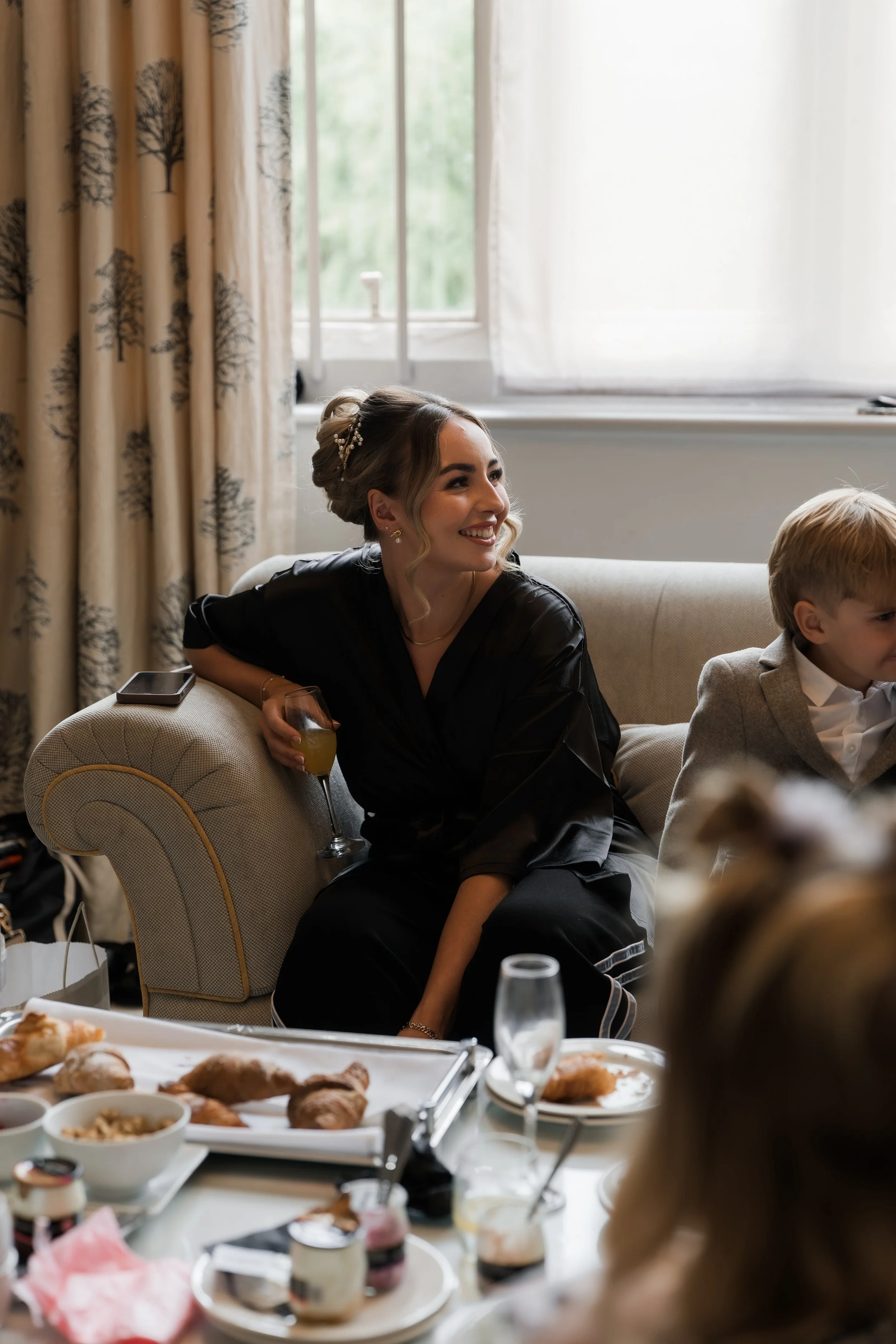 Woman in black dress sitting on a beige sofa at a social gathering, holding a drink, with a young boy in a gray blazer nearby, surrounded by food and drinks on a table.