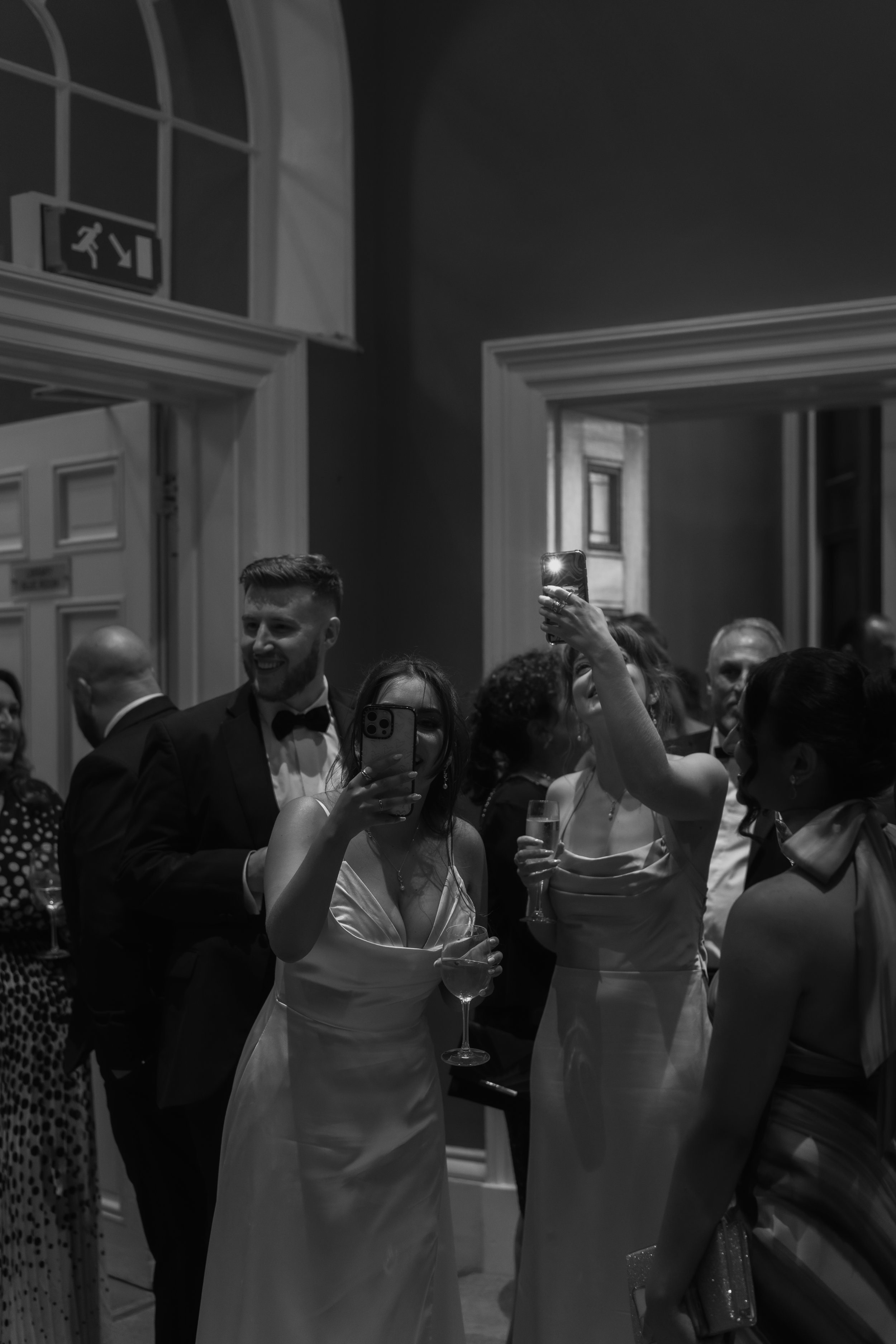 Group of people at a formal event taking photos with smartphones, some holding glasses of wine, in an elegant room with high ceilings and large windows.