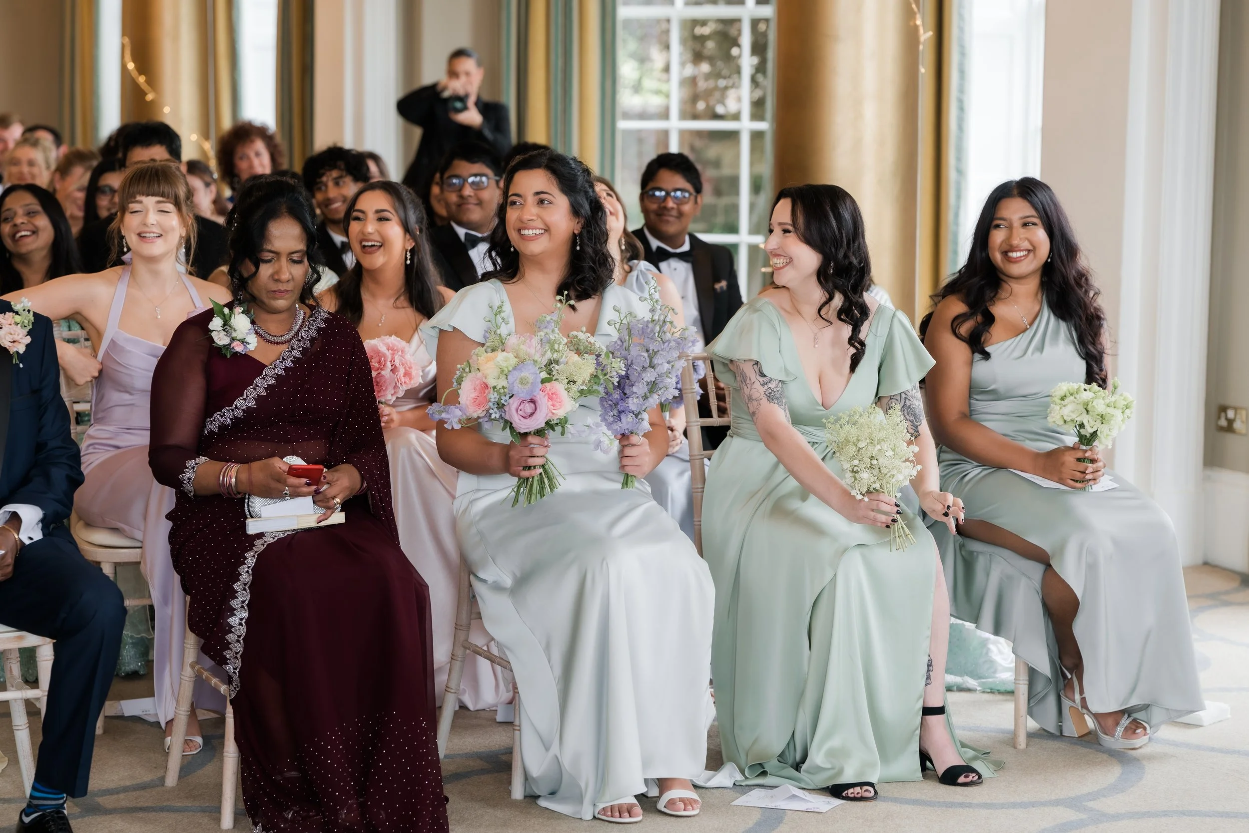Group of diverse women and men in formal wedding attire sitting in a decorated room, smiling, with some holding bouquets of flowers, during a wedding ceremony.