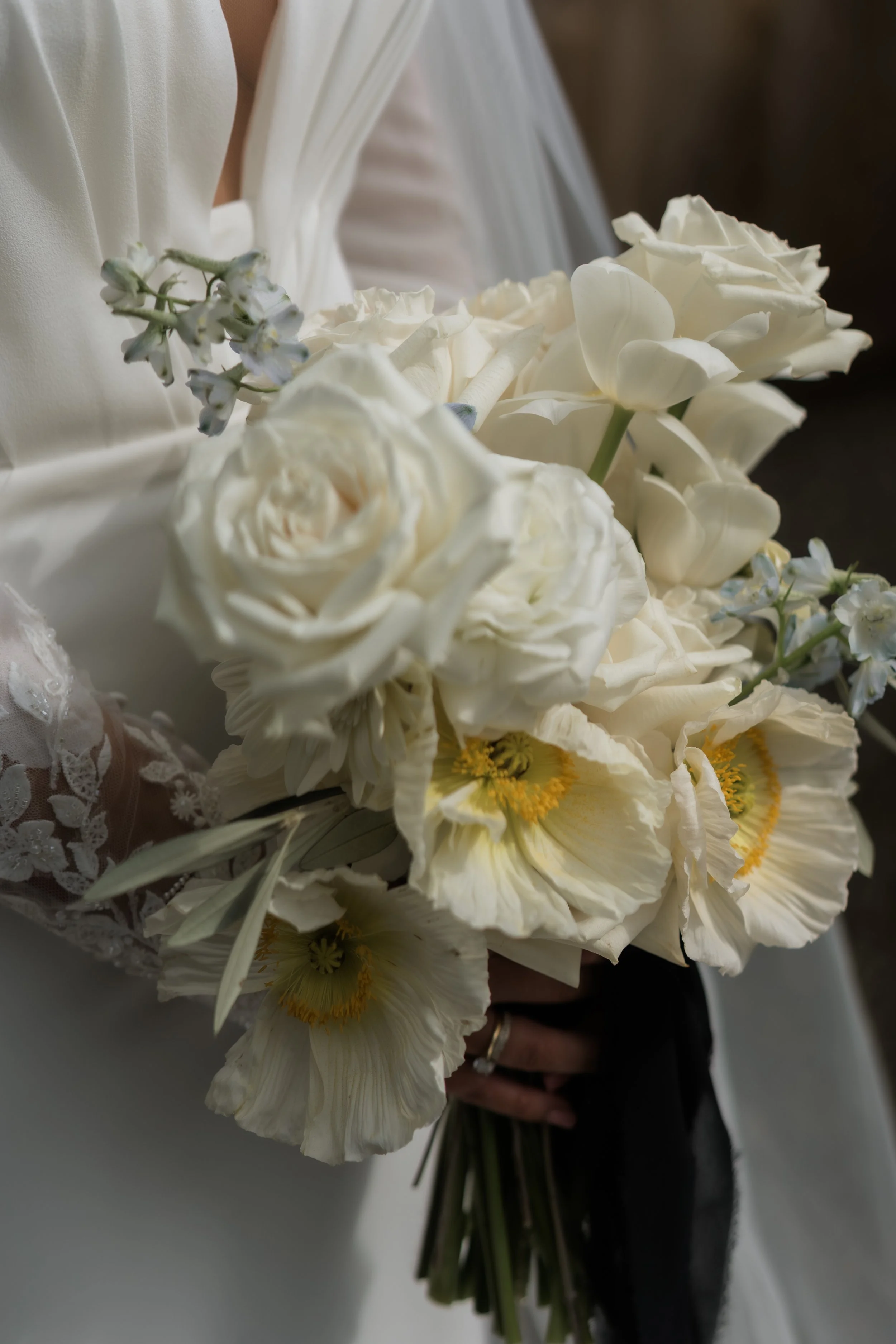 Close-up of a bride holding a bouquet of white flowers, including roses, tulips, and other blooms, with lace detail on her dress sleeve.