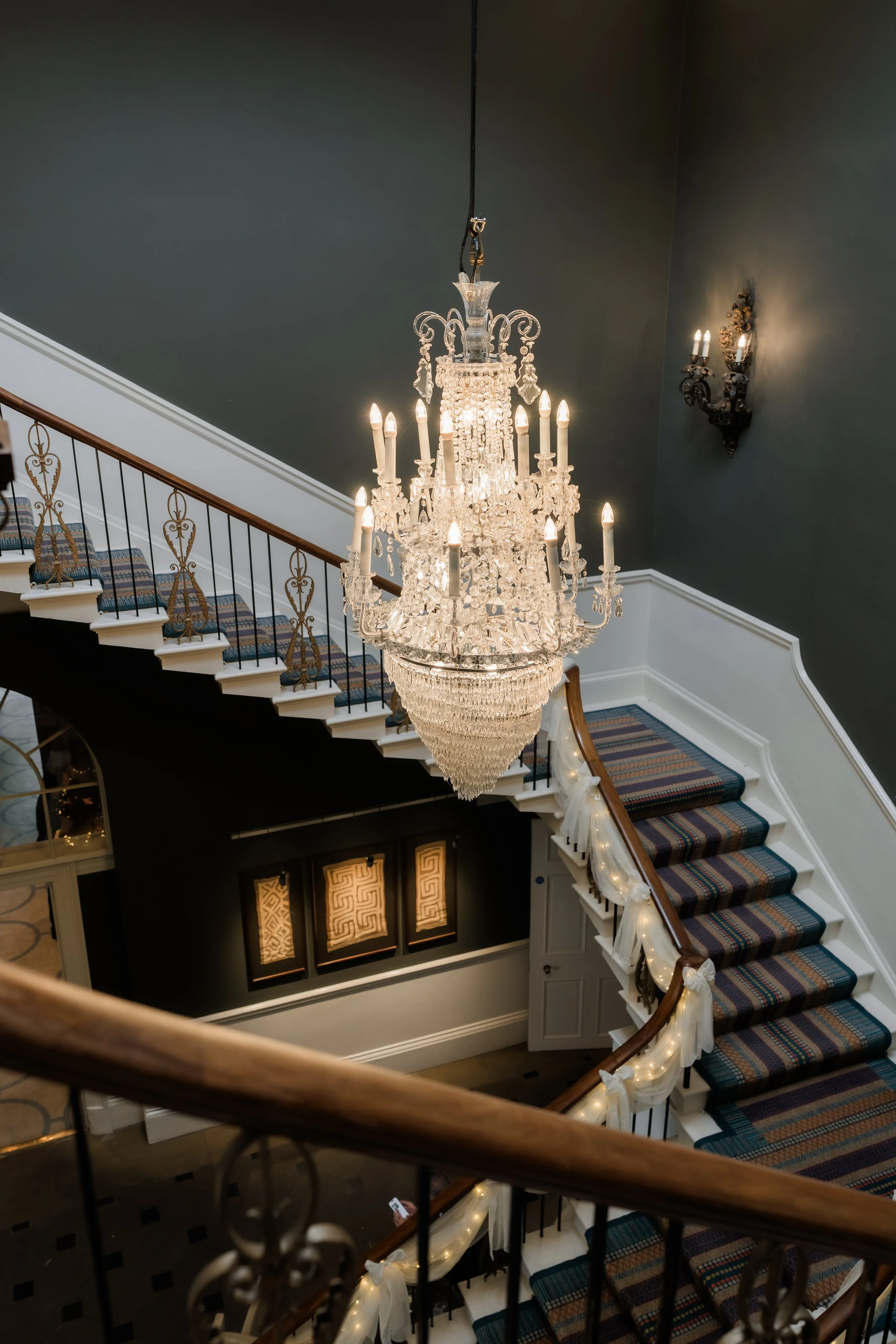 A grand chandelier hanging in a staircase hallway with dark walls, a wall sconce, and decorated staircase railings.