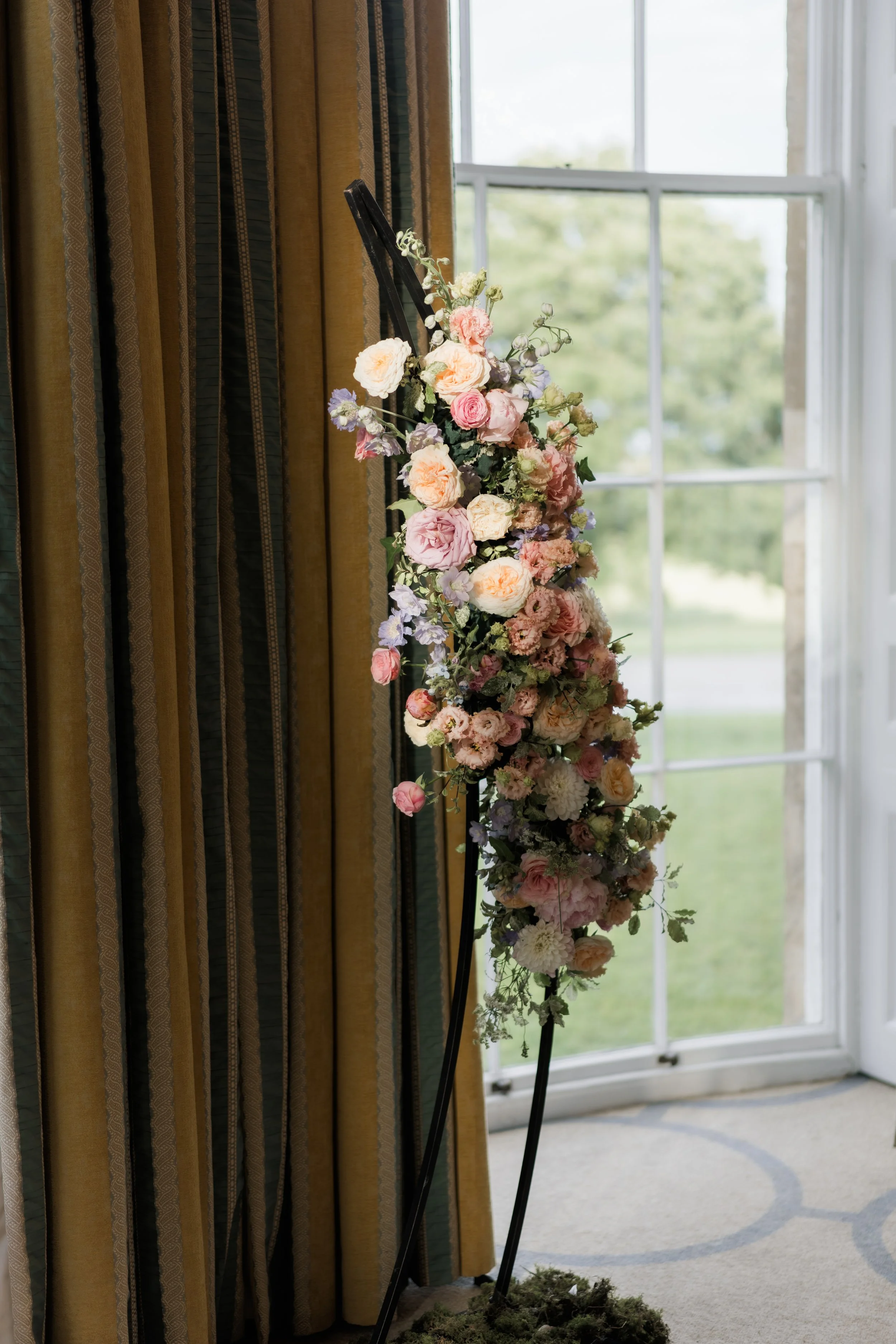 A flower arrangement on a black metal easel, placed indoors near a window with striped curtains and a view of the outdoors.