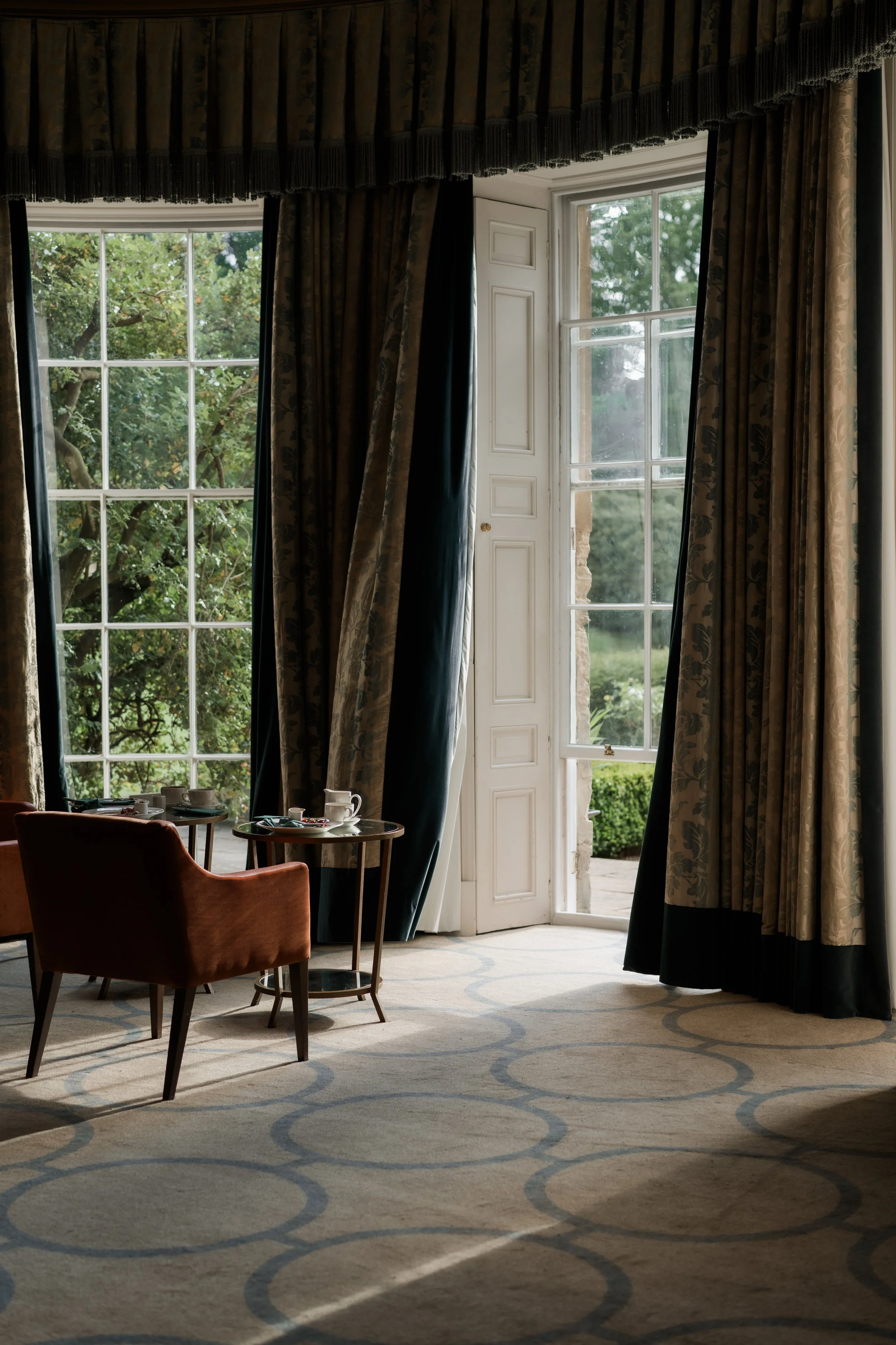 Interior of a room with large windows and a door opening to a garden, with elegant curtains and a small table with tea cups and chairs.
