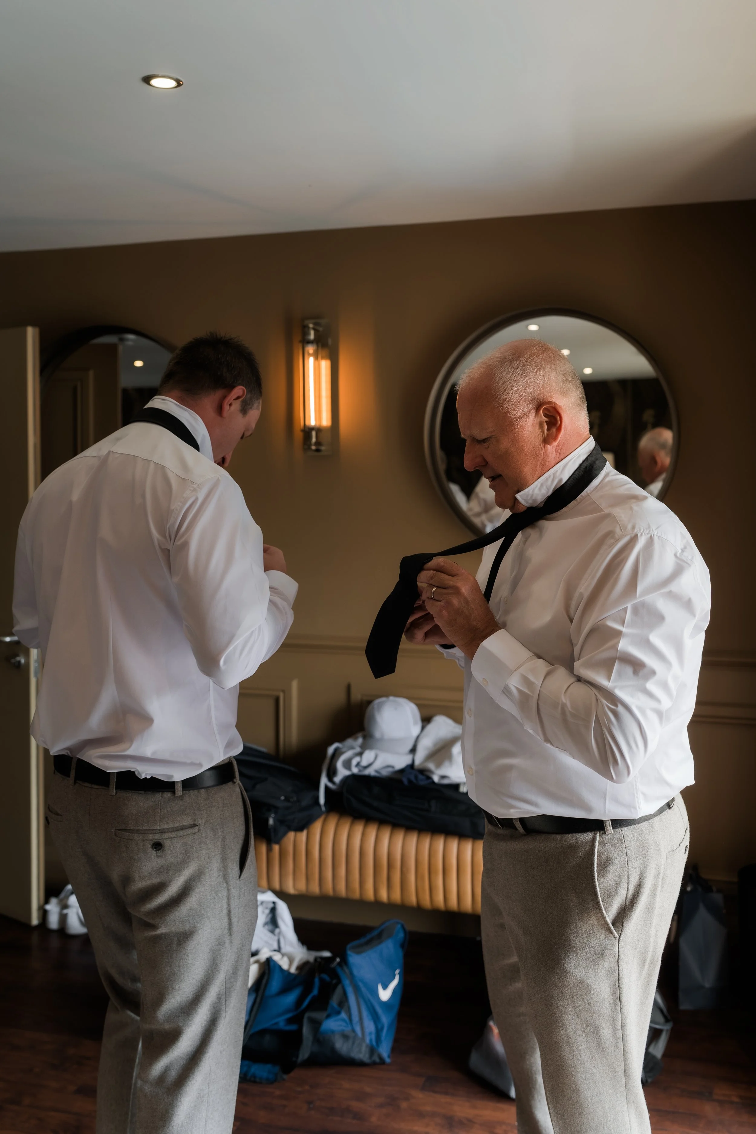 Two men dressing in formal white shirts and beige trousers are in a room with warm lighting, preparing for an event. The room has a bench with clothing and bags, and a round mirror on the wall.