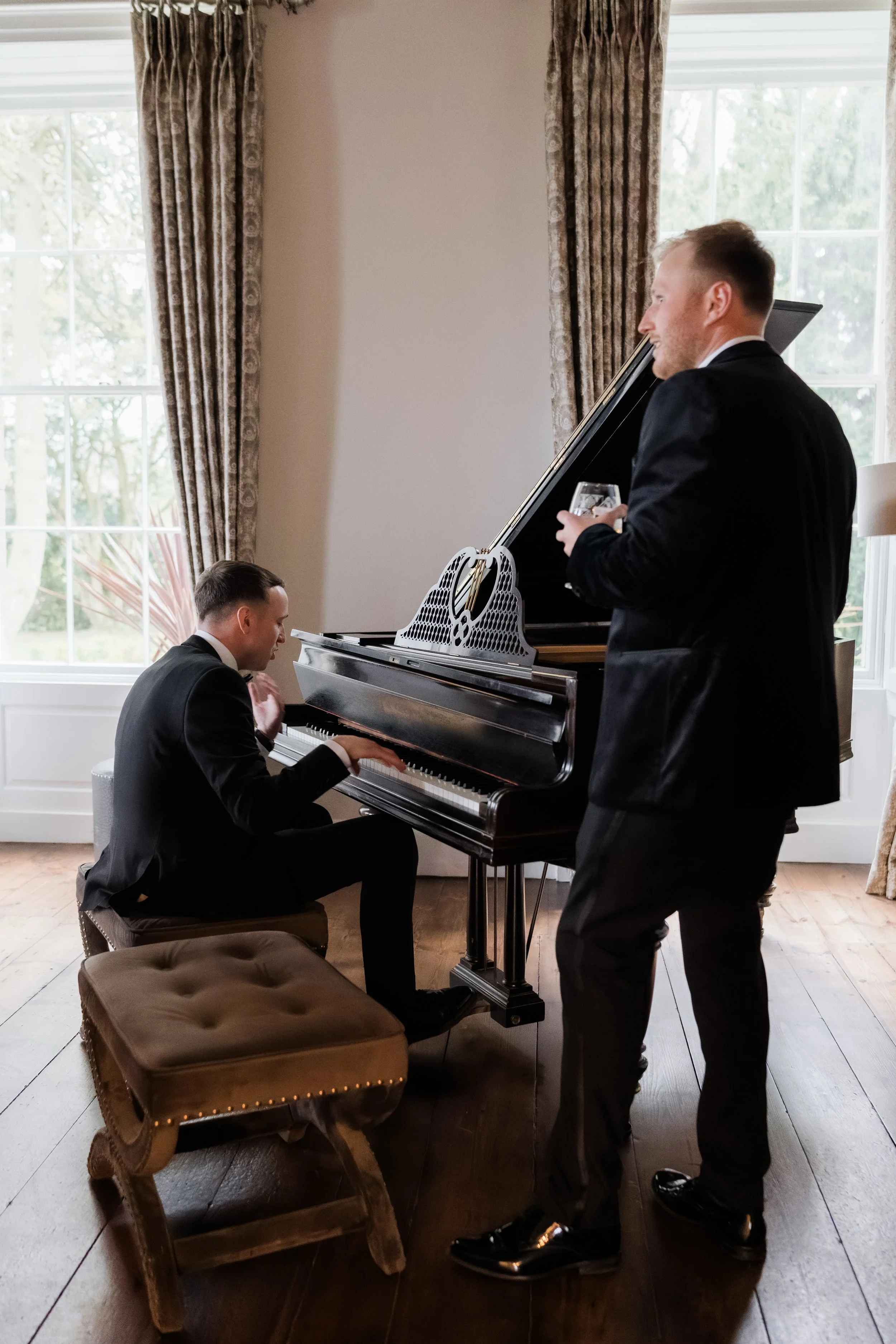 Two men in formal suits are in a room with large windows and curtains, one playing a grand piano and the other standing with a glass in hand.