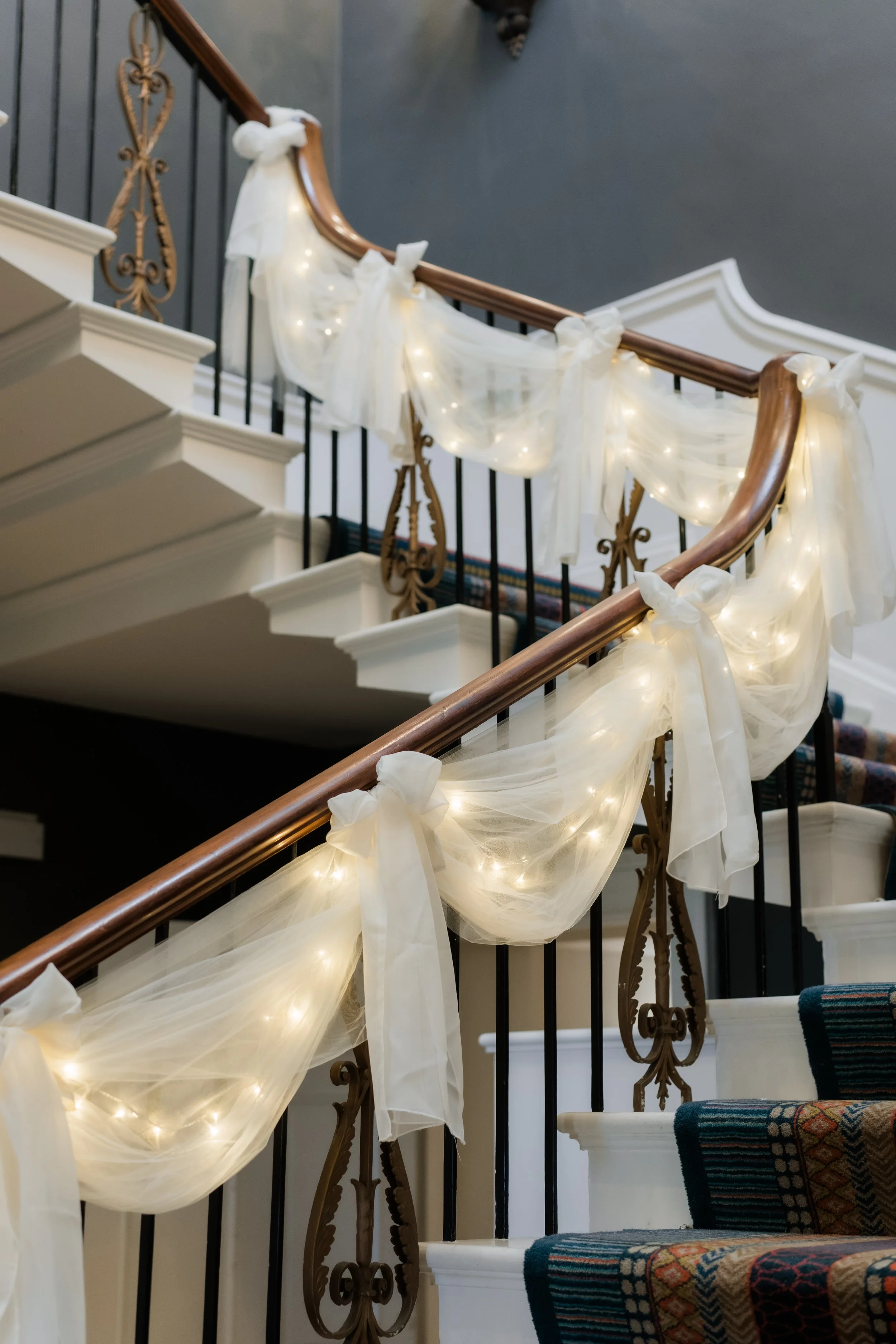 Staircase decorated with white fabric and string lights, tied with bows, in an elegant indoor setting.