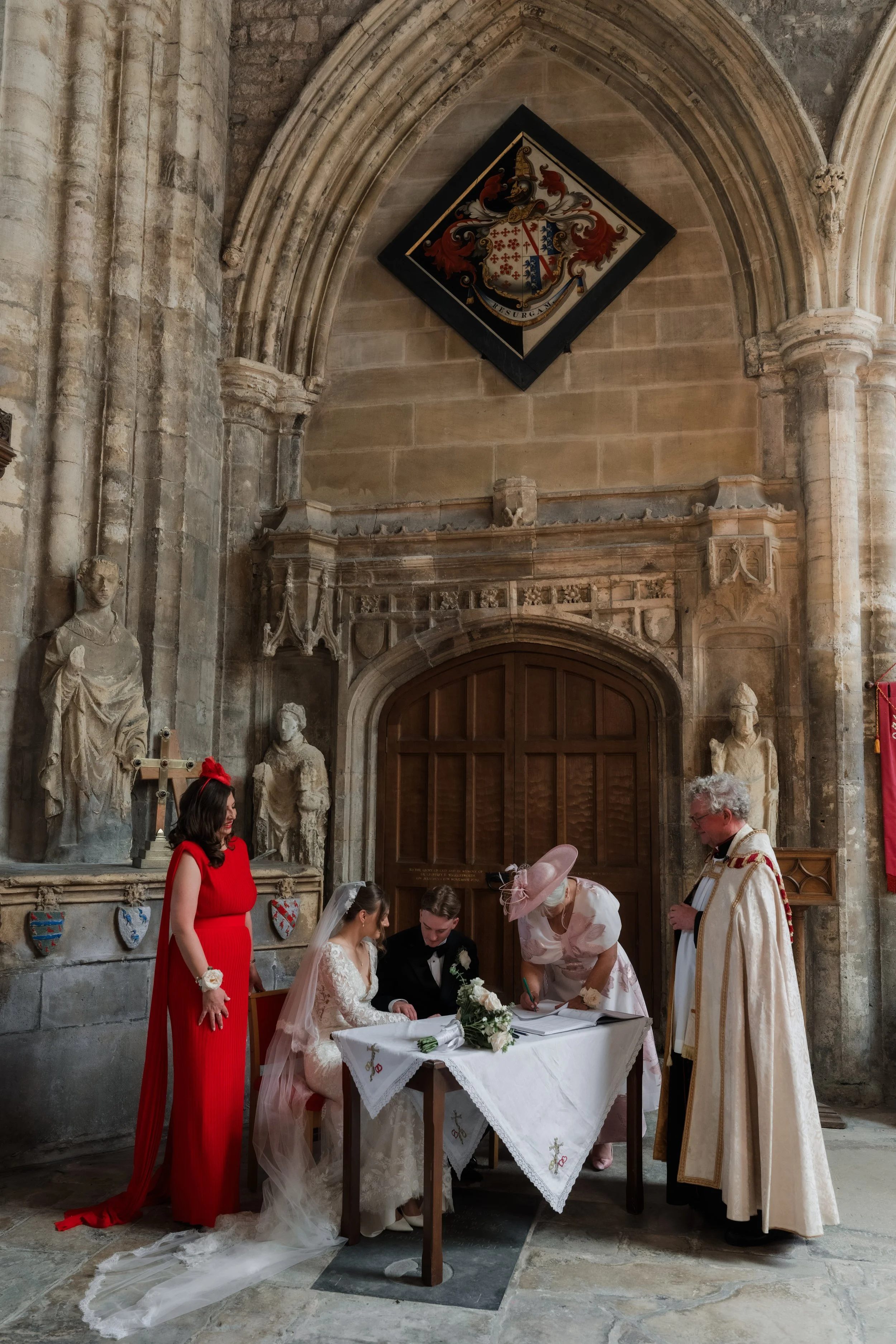 A wedding ceremony taking place inside a church with stone walls and statues. The bride and groom are seated at a table, signing documents, with a woman in a pink hat and a woman in a red dress standing nearby. A priest in ceremonial robes is present