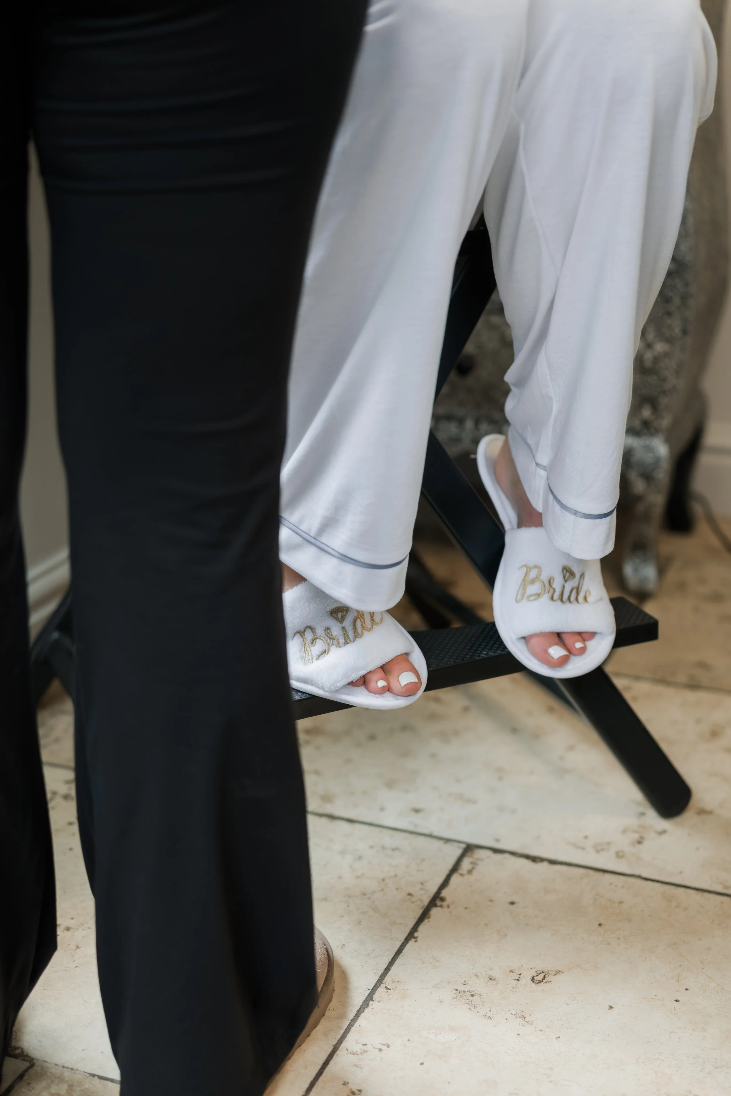 A bride wearing white slippers with 'Bride' embroidered in gold, sitting on a black chair.