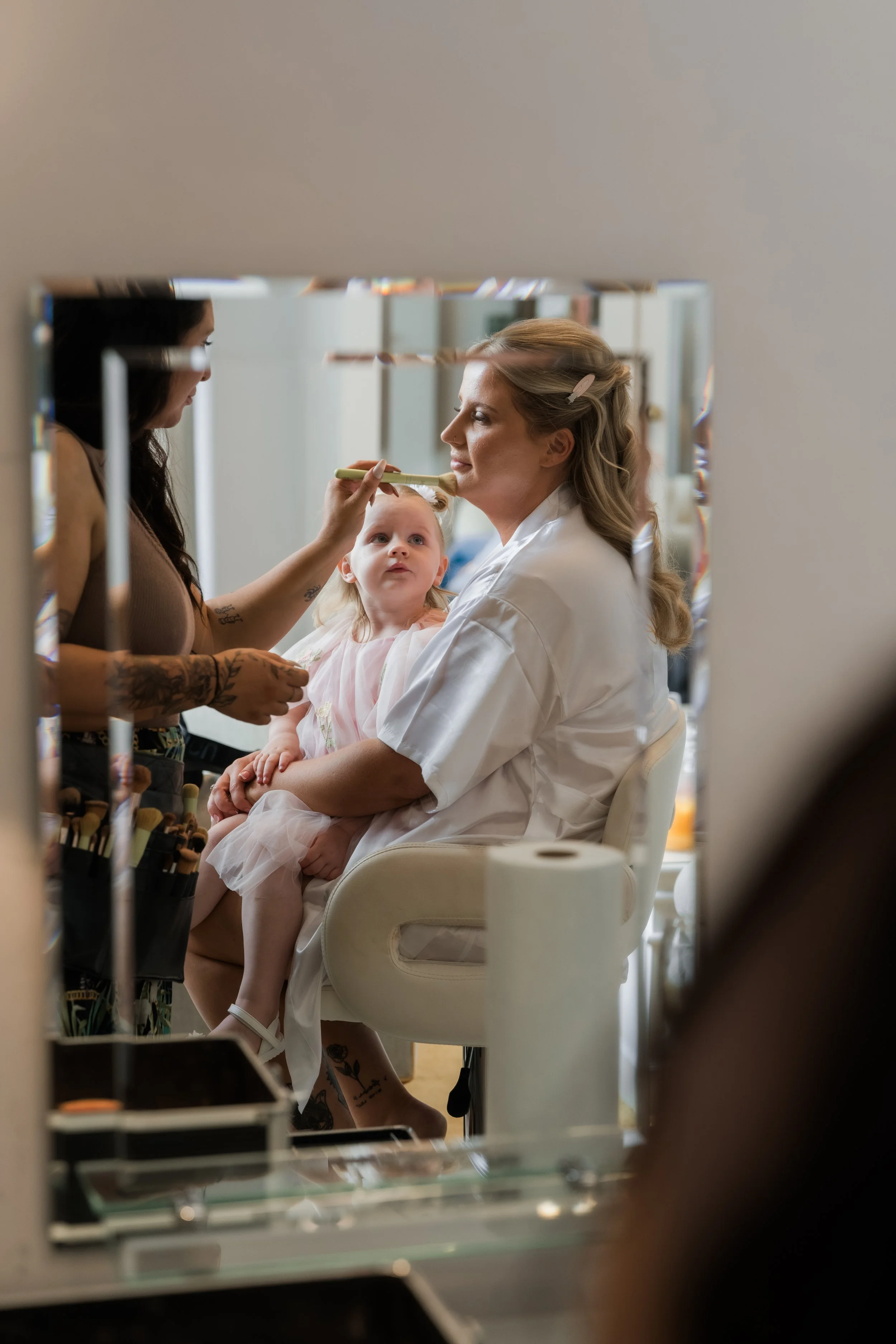 L&J Wedding Photography & Videography. Documentary Yorkshire Weddings. Bride having make up done while little girl sits on knee and looks up at her. Reflection taking in mirror