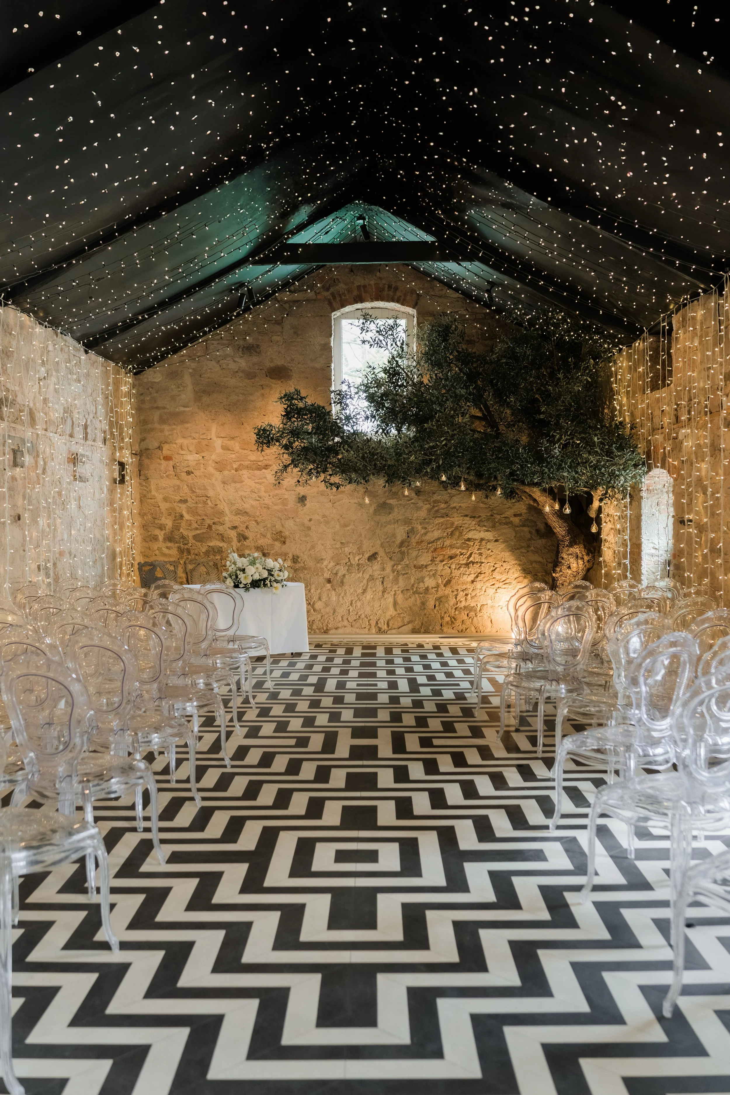 An indoor event space with a black and white geometric patterned floor, a large tree on the stage area, string lights hanging from the ceiling, and a table with flowers at the front.