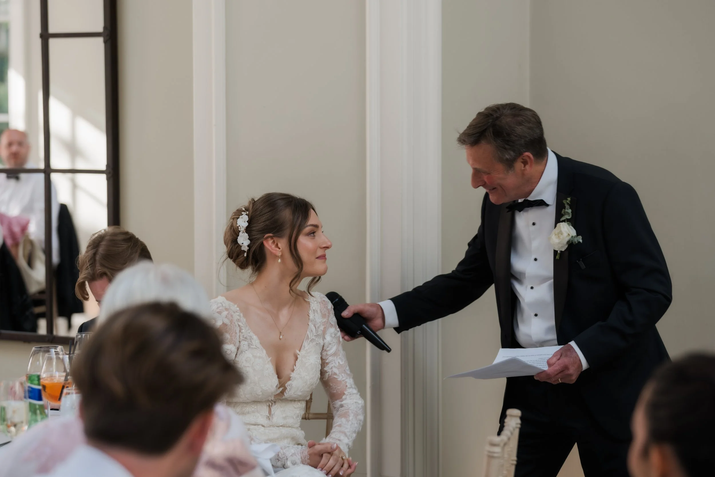 A bride with brown hair wearing a lace wedding dress and floral headpiece looks at a man in a tuxedo with a white boutonniere, who is holding a paper and a microphone, during a wedding ceremony.