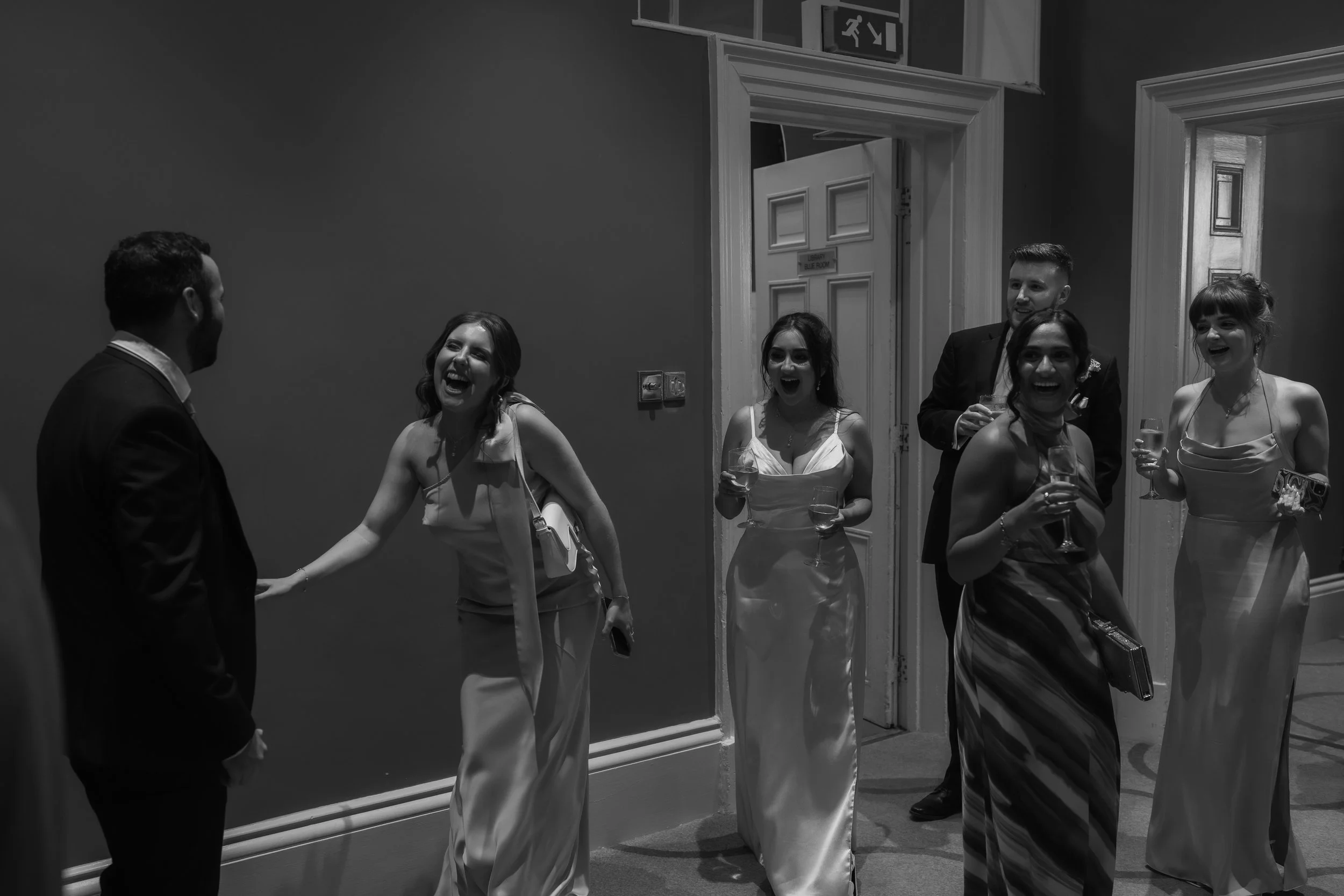 Six people, including three women and two men, are standing in a hallway, laughing and holding glasses of champagne, engaging in conversation.