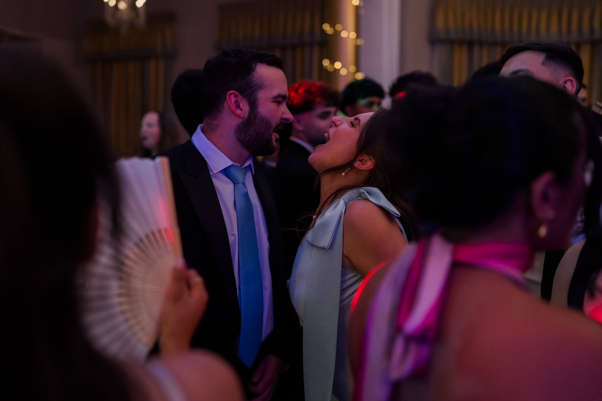 A man and woman are close to each other, appearing to be arguing or shouting, at a crowded indoor event with warm lighting and blurred background guests.