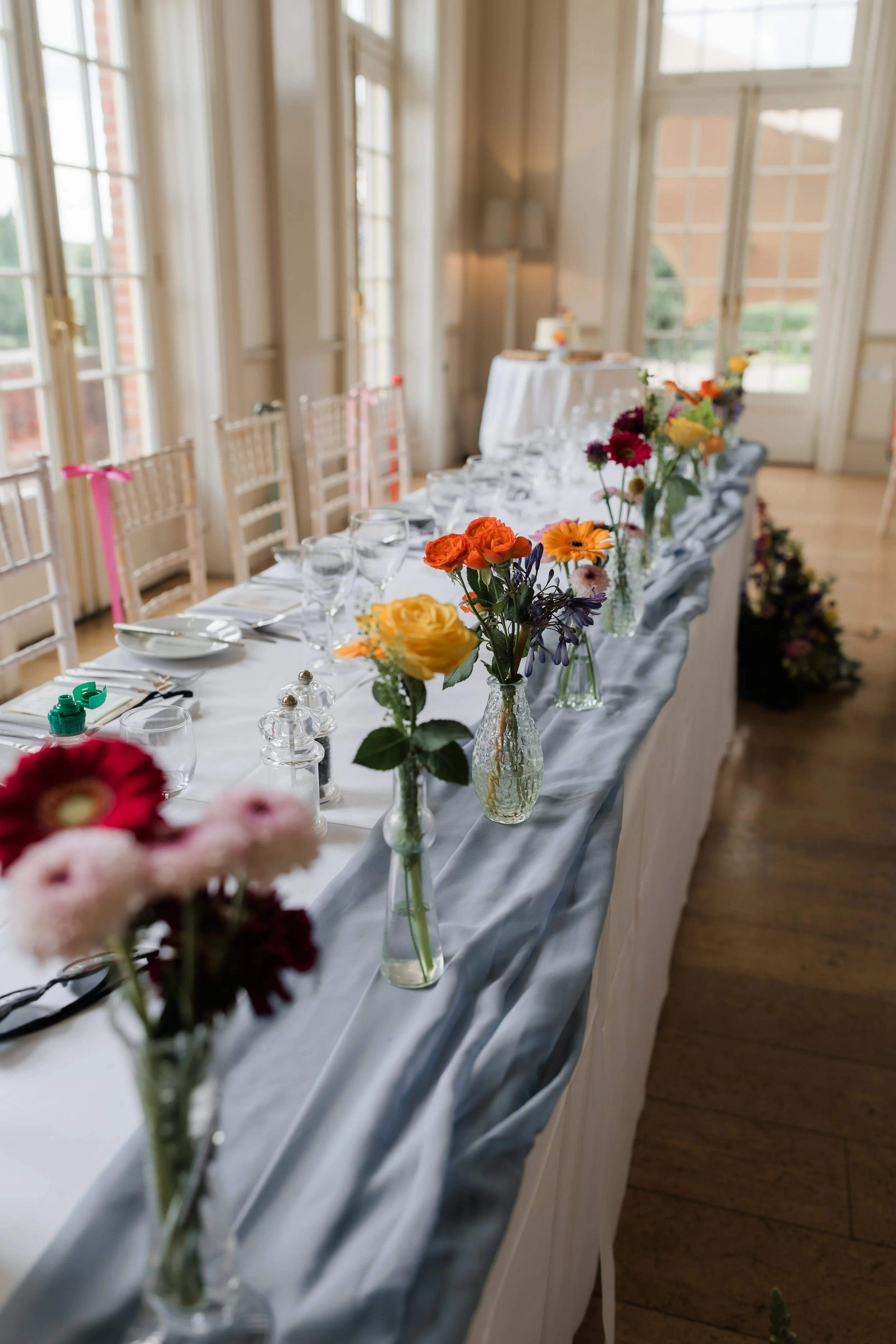 Elegant indoor dining table decorated with colorful flower arrangements in glass vases, set for a formal event with white tablecloths, glassware, and silverware, in a bright room with large windows.