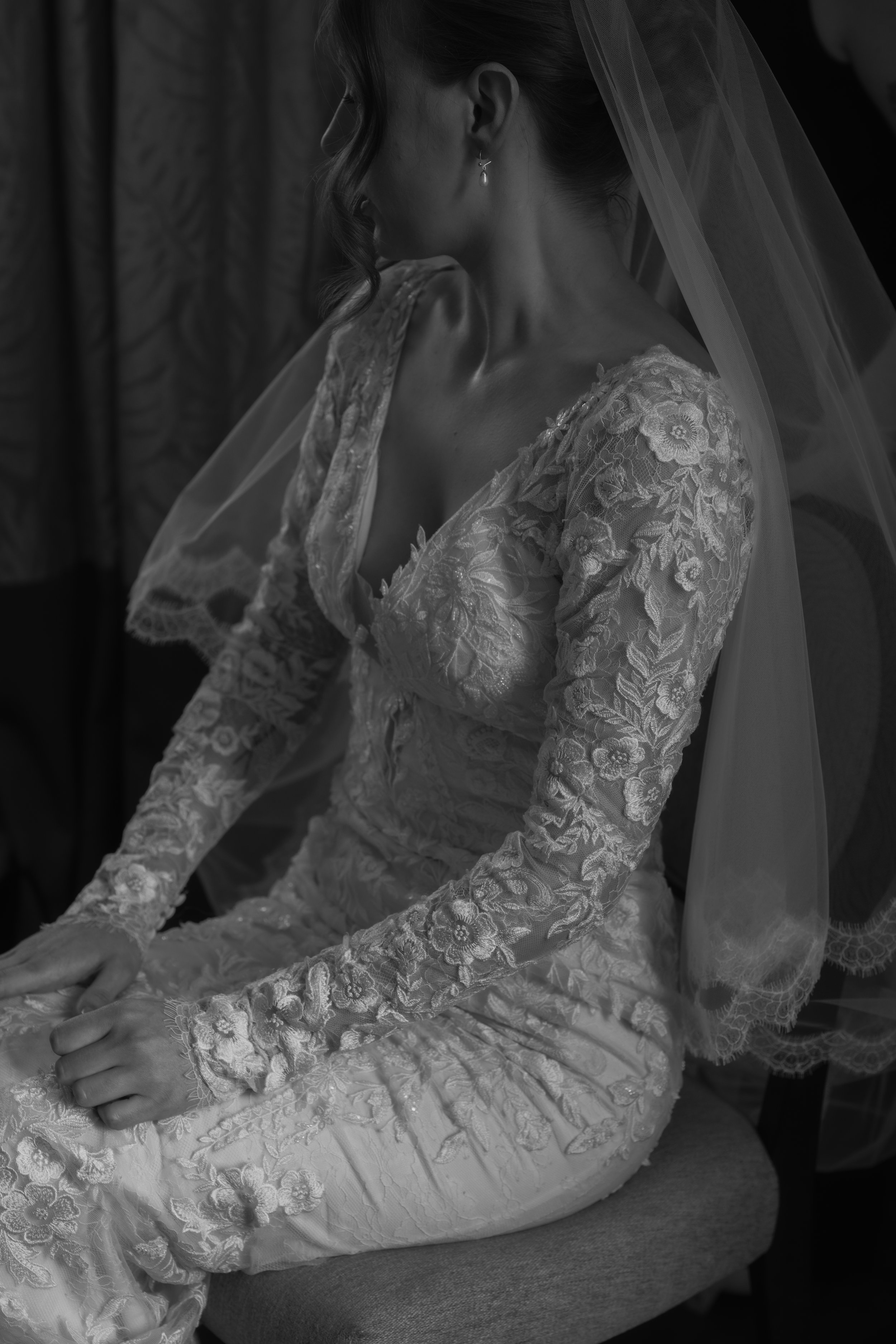 Black and white photograph of a bride sitting, wearing an embroidered wedding dress with floral patterns, lace sleeves, and a veil. She is looking down with her hands folded on her lap.