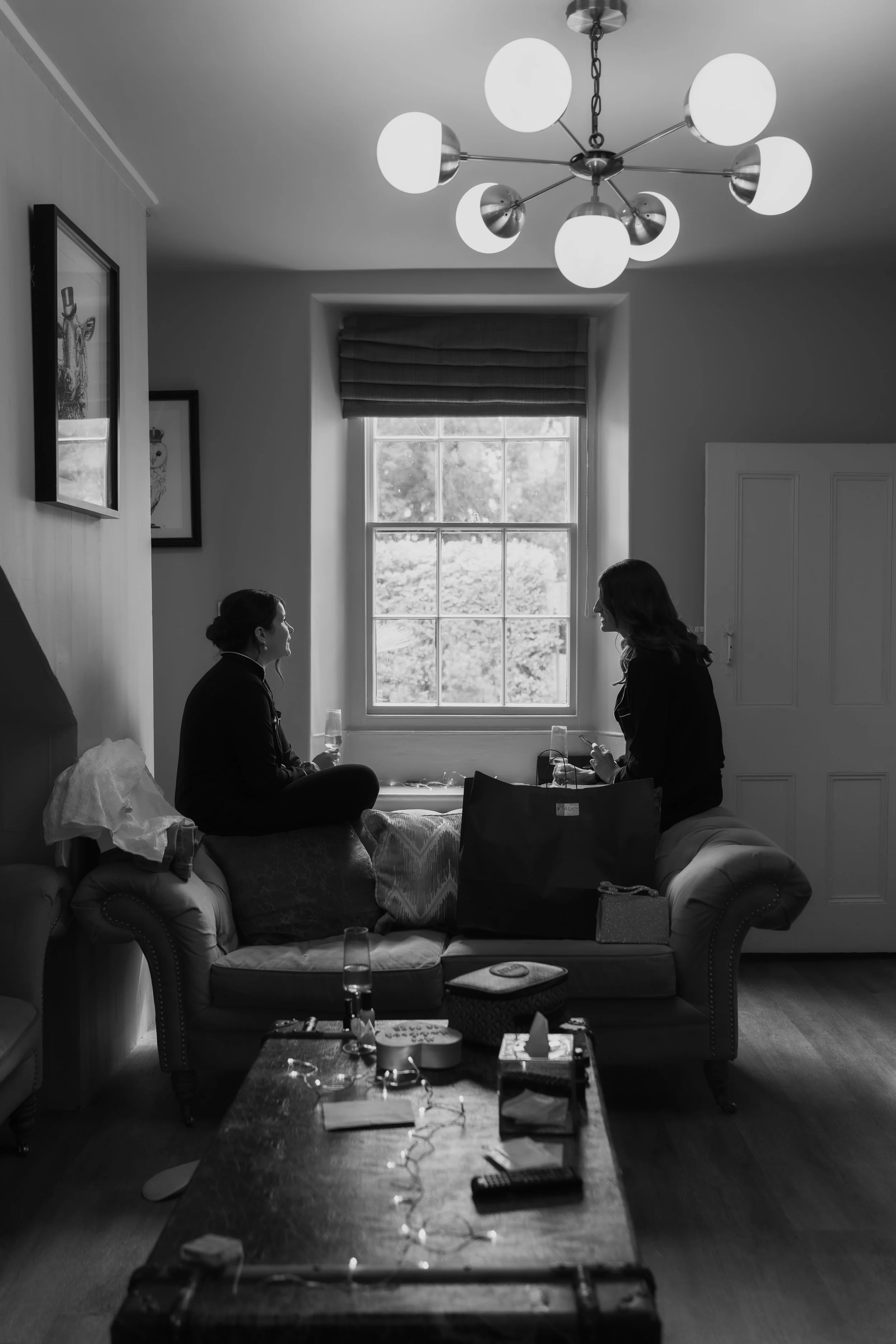 Two women sitting on a window ledge, talking and holding drinks in a living room with a modern chandelier and framed art on the wall.