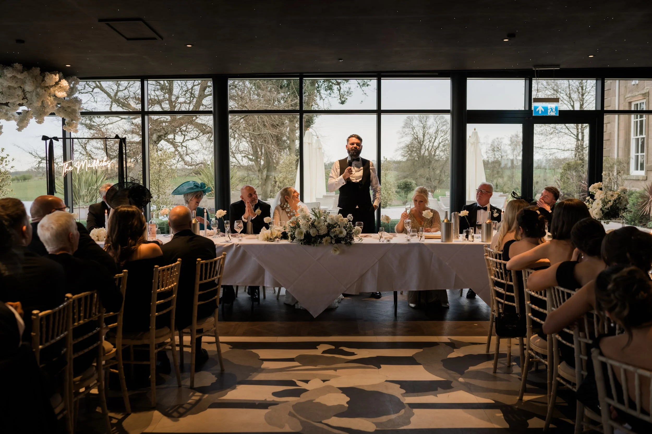 A wedding reception with guests seated around a long table. A man is standing and speaking into a microphone at the head of the table, with a floral arrangement in front. Large windows show an outdoor landscape with trees and fields behind the scene.