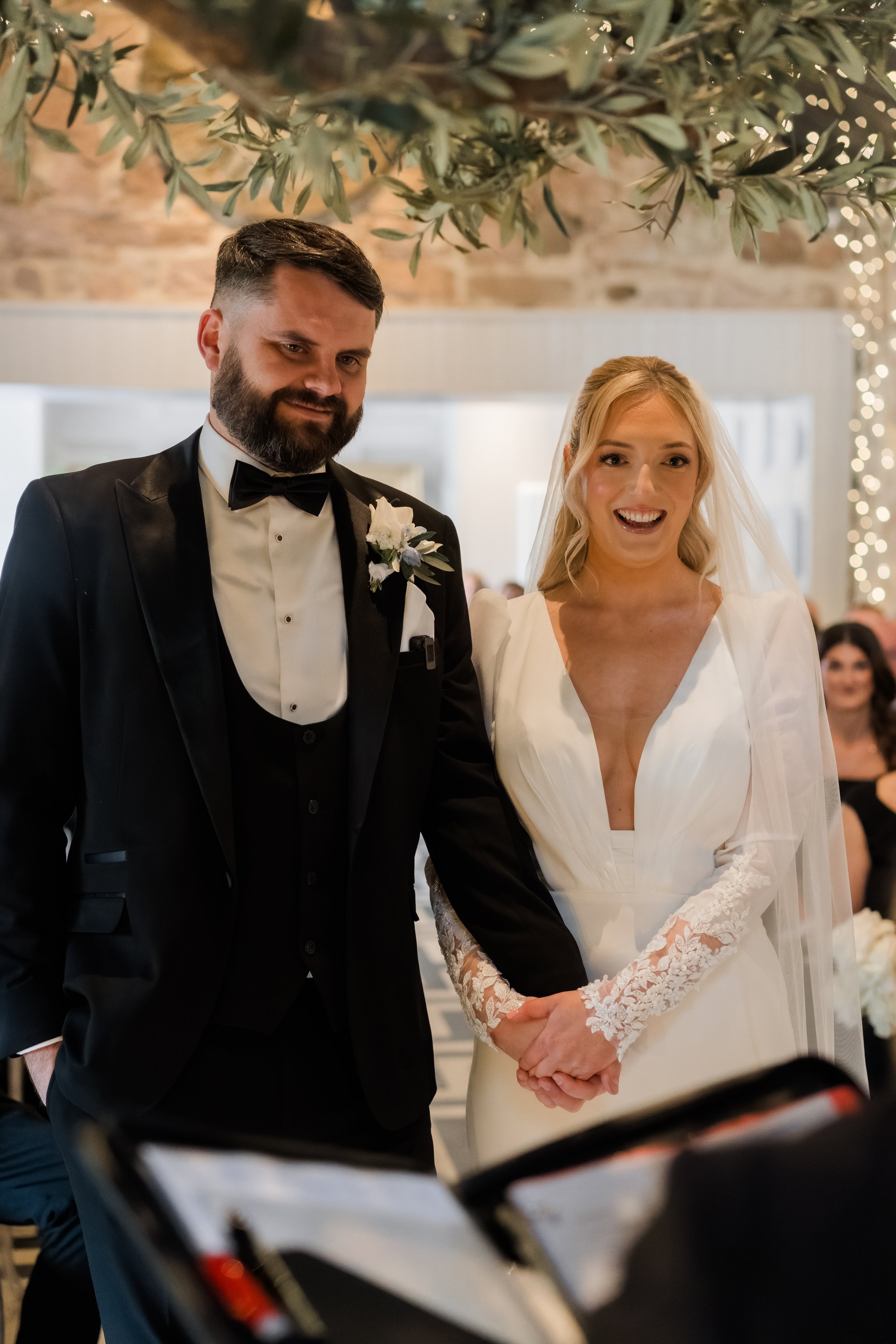 A bride and groom holding hands during a wedding ceremony, with the bride smiling and the groom looking serious, in an indoor decorated venue.