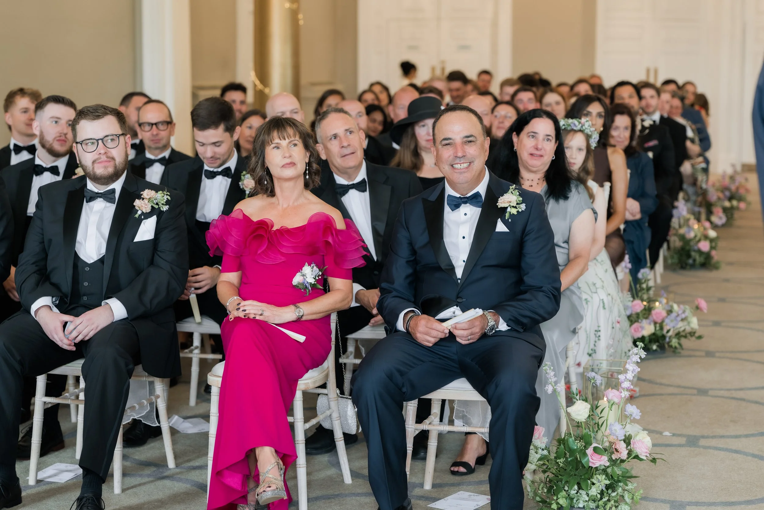 A group of wedding guests seated, dressed in formal attire, in a decorated indoor venue.
