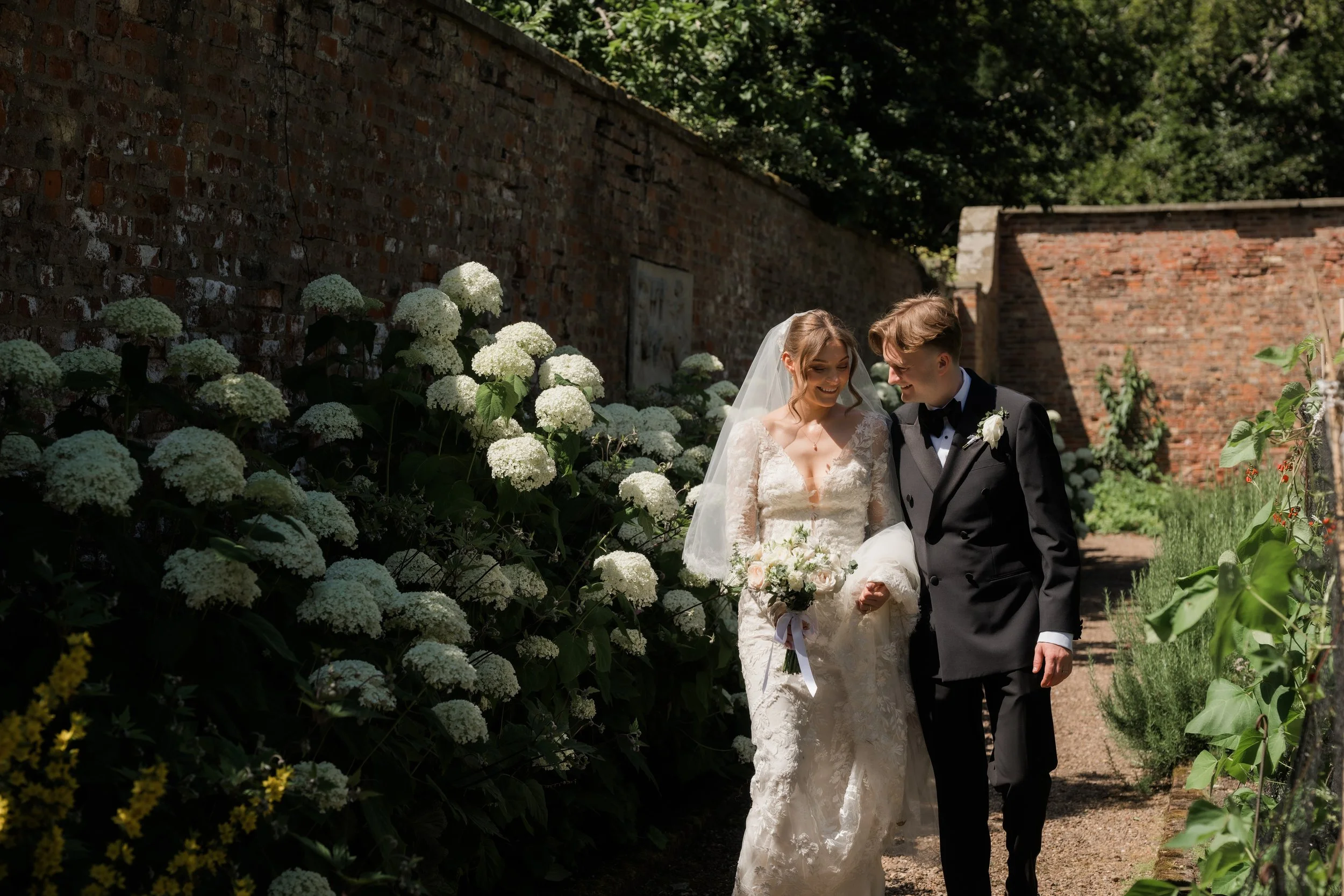 Bride and groom walk past white hydrangea border smiling at each other 