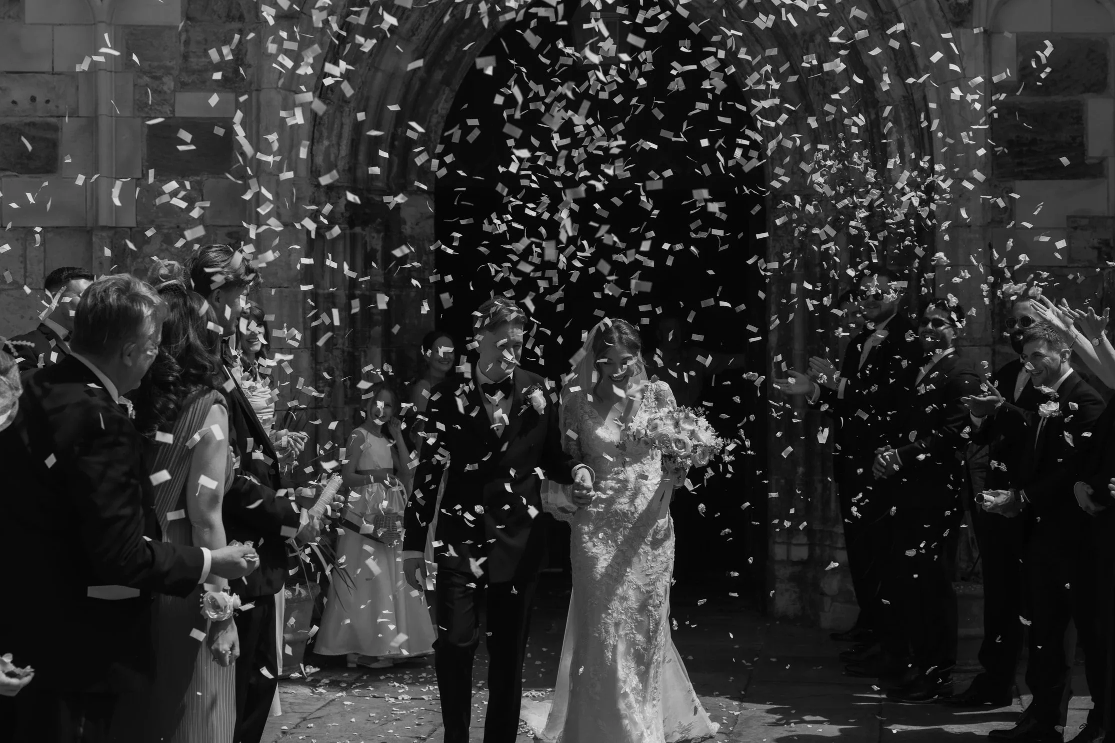 Wedding celebration with confetti falling around a bride and groom among friends and family, standing outside a stone building with an archway.