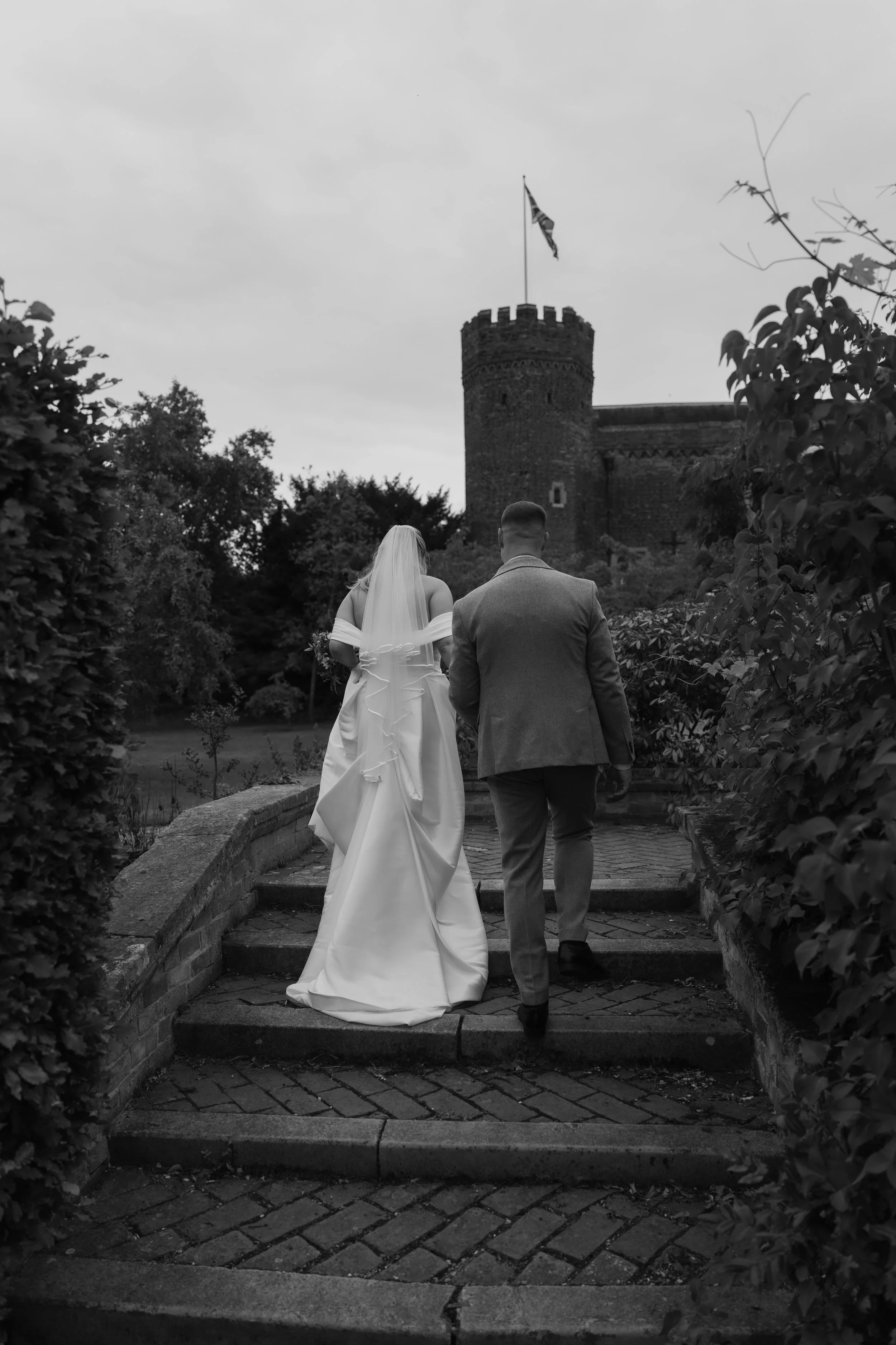 A bride and groom walk up stone steps towards a castle with a tower and an American flag, surrounded by bushes and trees.