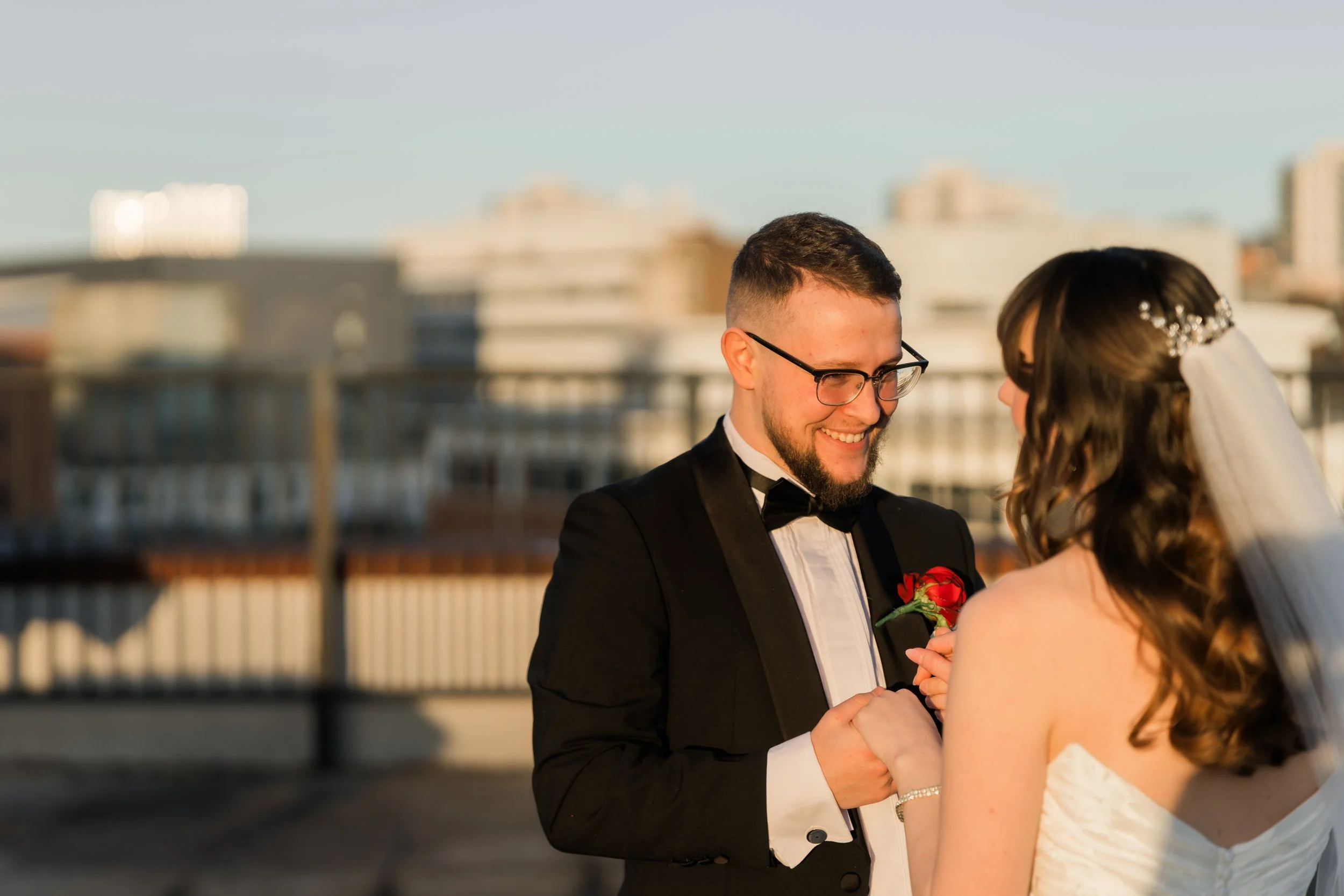 A newlywed couple holding hands and smiling at each other on a rooftop during sunset, with an urban skyline in the background.