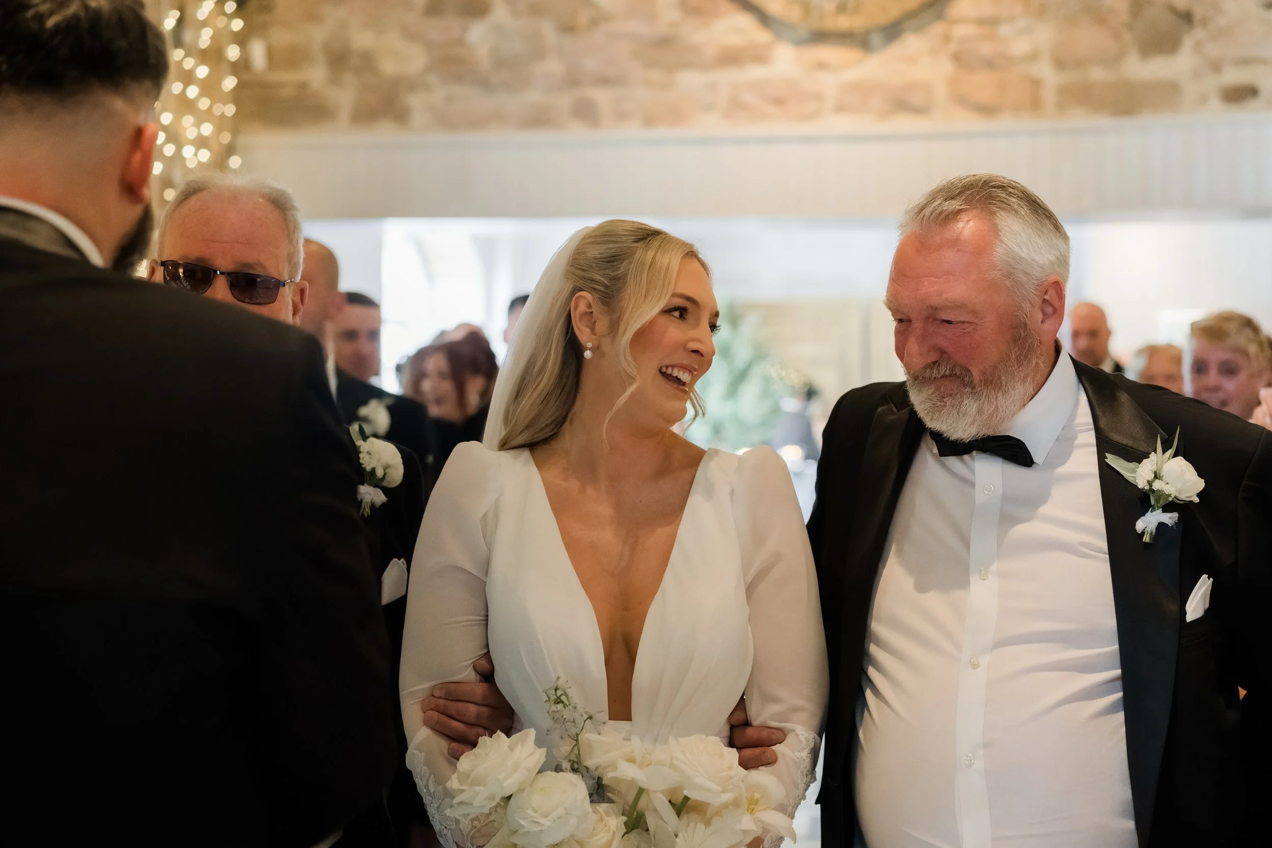 Bride in white wedding dress smiling and talking with an older man in a tuxedo at a wedding reception.