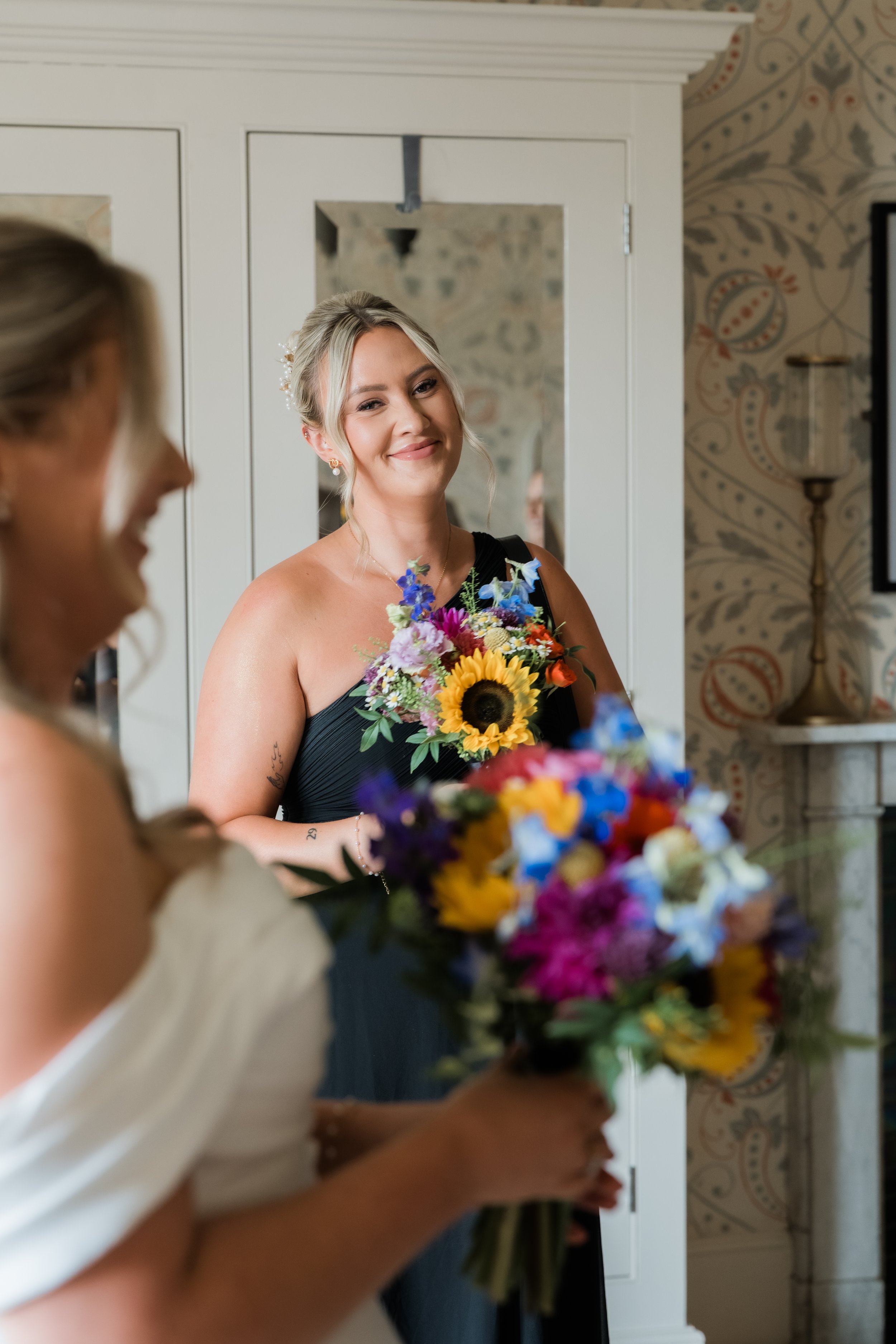 A woman in a black dress smiling and holding a colorful bouquet of flowers, with another woman in a white dress and holding a bouquet in the foreground.